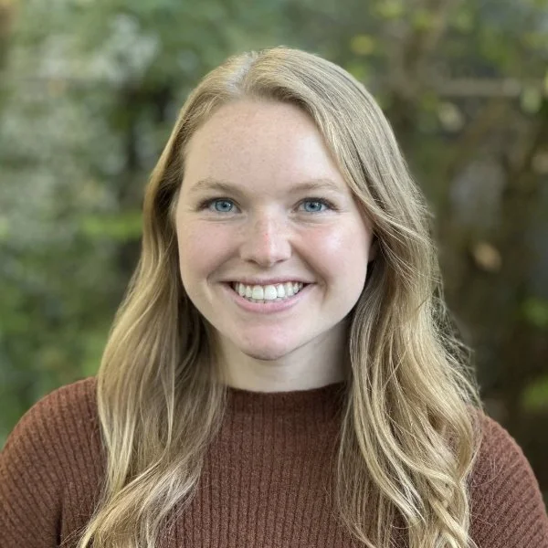 Therapist Jocelyn Peters, wearing a brown sweater, smiling outdoors with blurred green foliage in the background.
