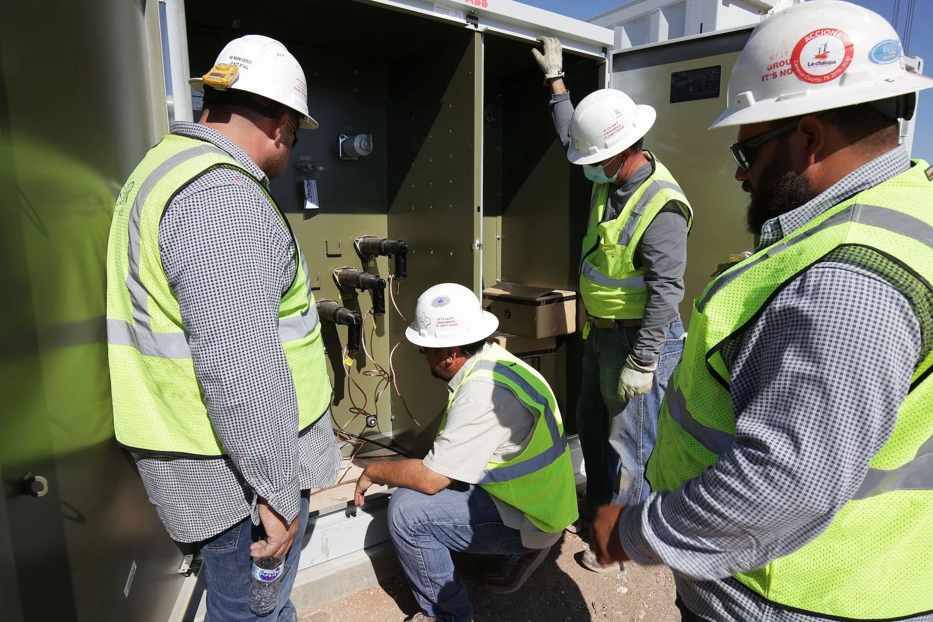 Group of five workers wearing safety glasses, helmets, and yellow safety vests working on electrical equipment outdoors.