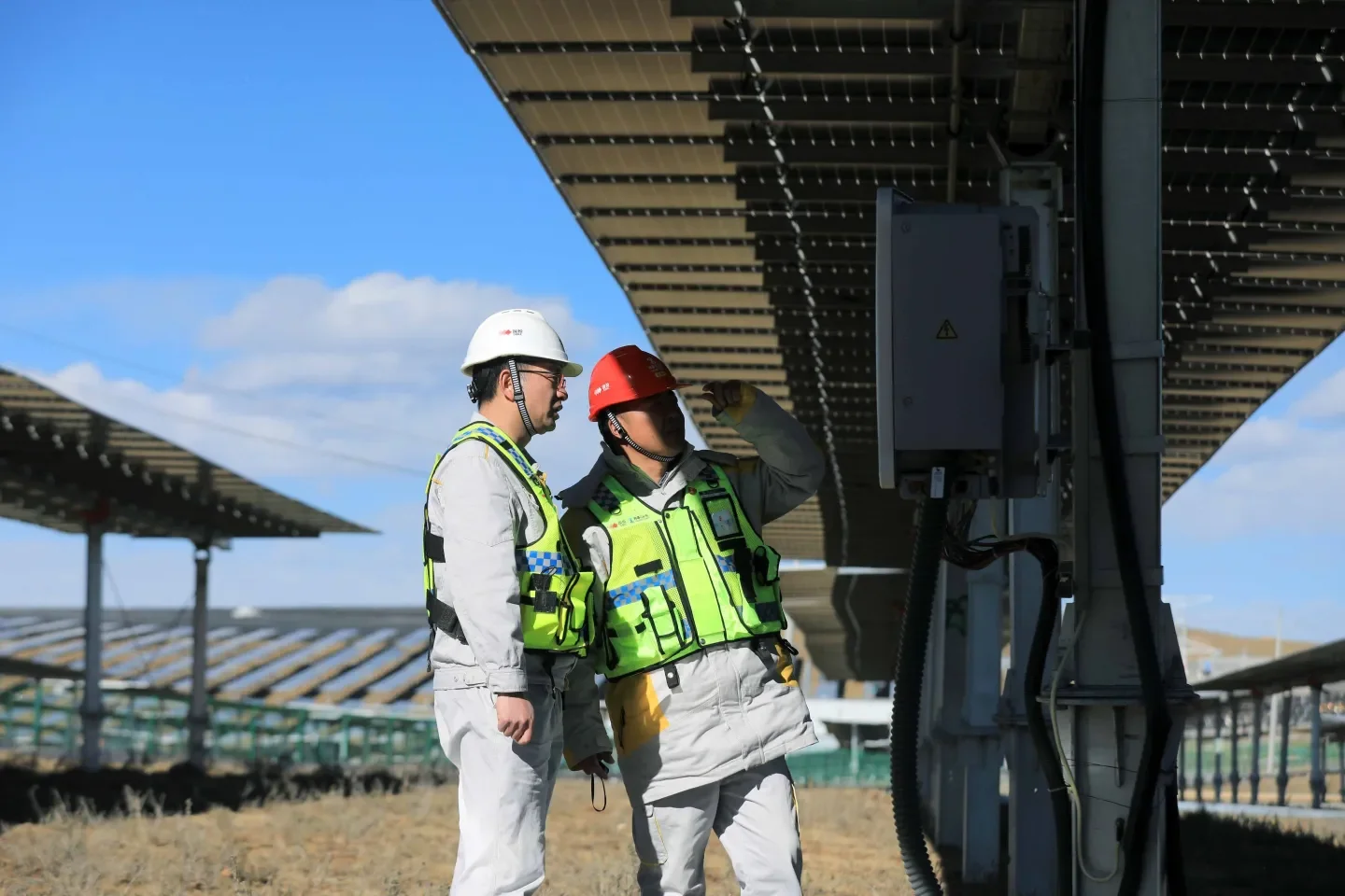 Two workers in safety gear, including helmets and vests, inspecting solar panels on a large solar farm under a blue sky.