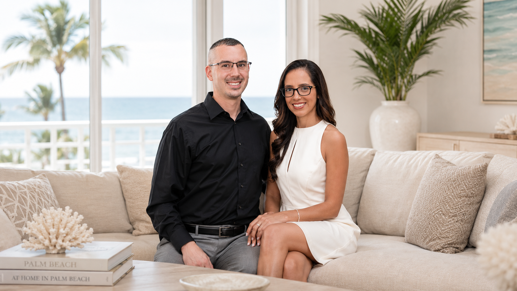 A couple (Michael and Pamela) sitting on a beige sofa in a bright living room with ocean view, palm trees outside, and decorative pillows, with a large potted plant in the background.