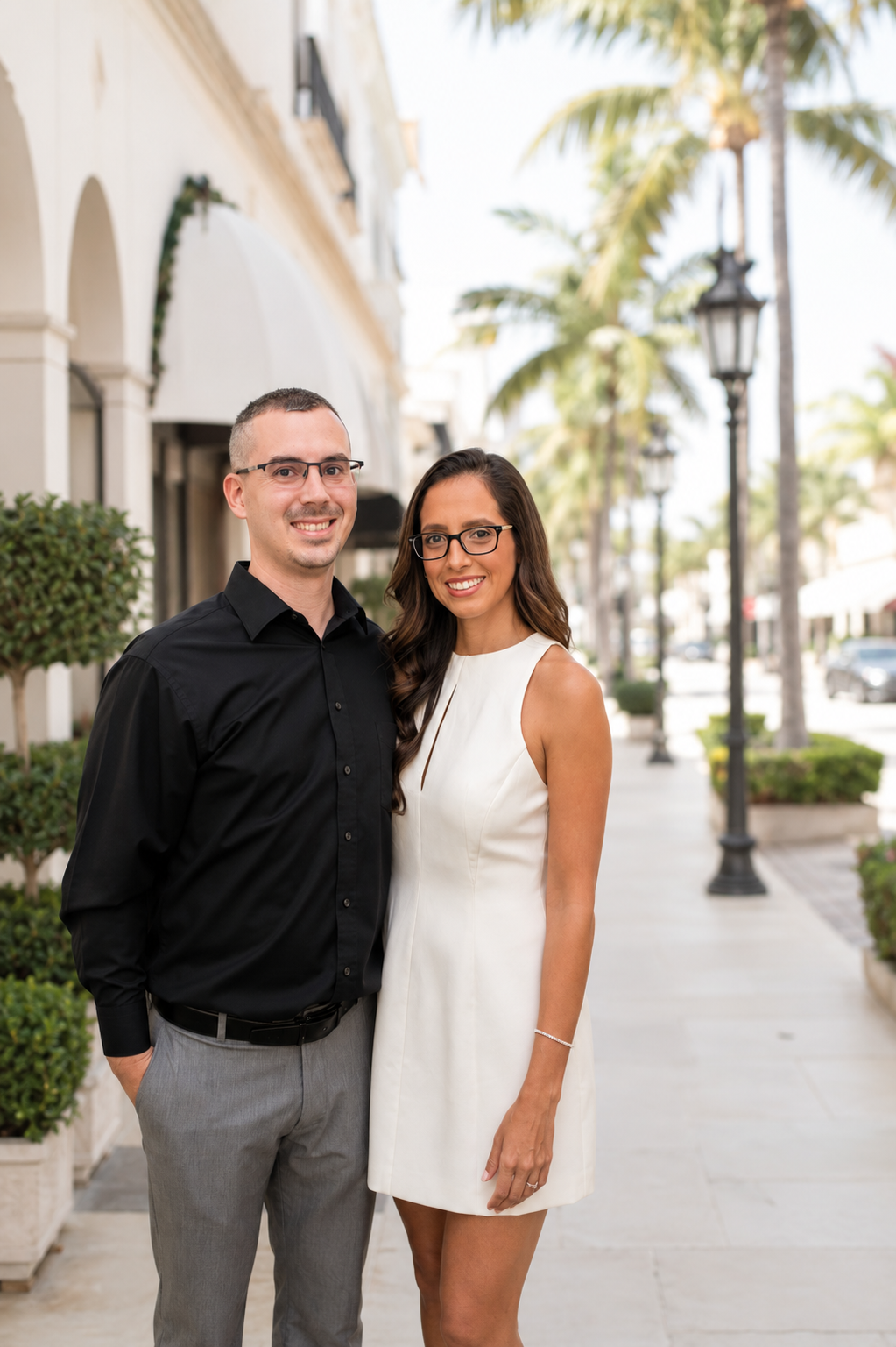 A smiling man (Michael) and woman (Pamela) standing together on a sunny Palm Beach sidewalk lined with palm trees, black street lamps, and buildings, enjoying a warm day.