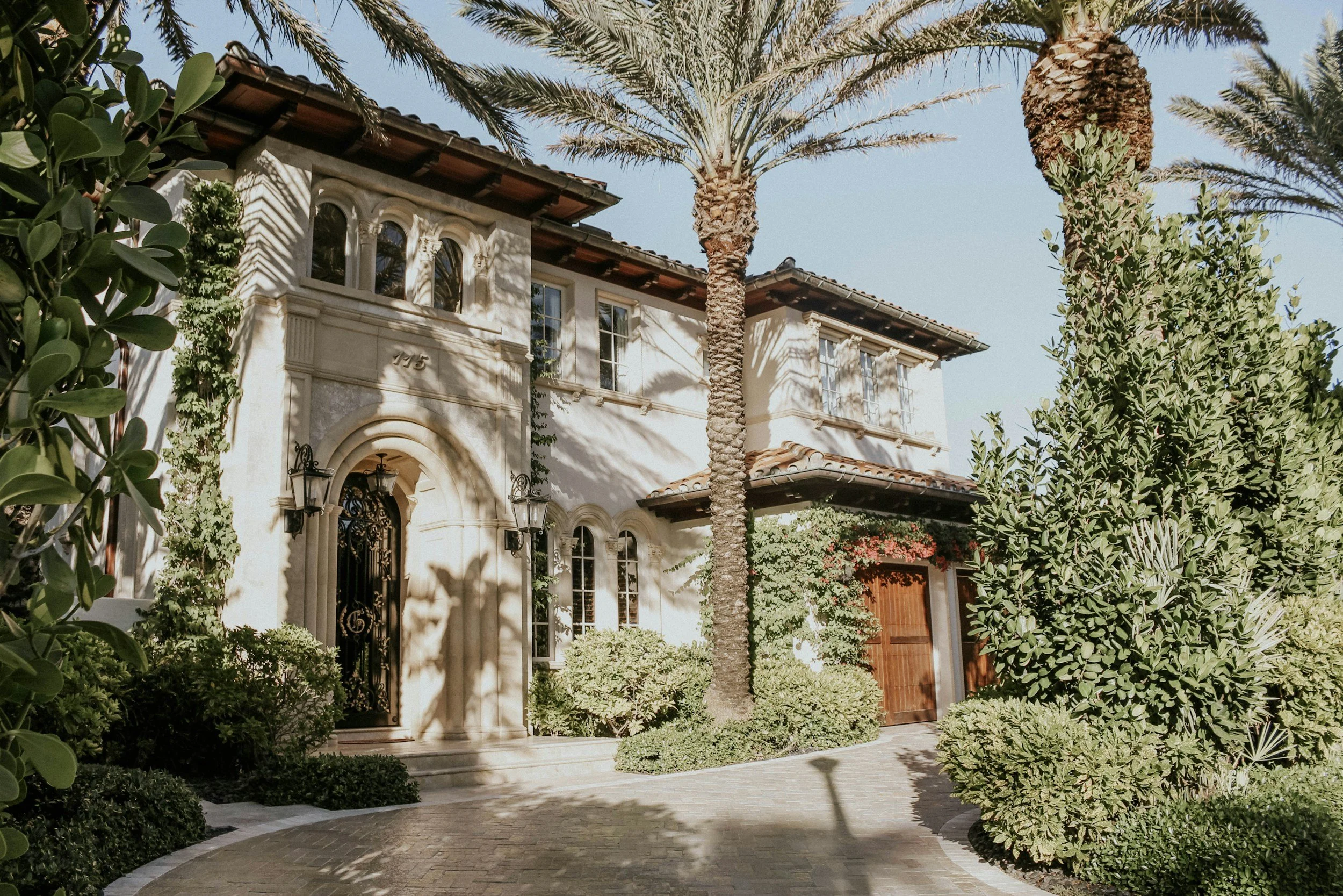 Luxury Palm Beach house with lush greenery, palm trees, and a stone driveway in a warm climate.