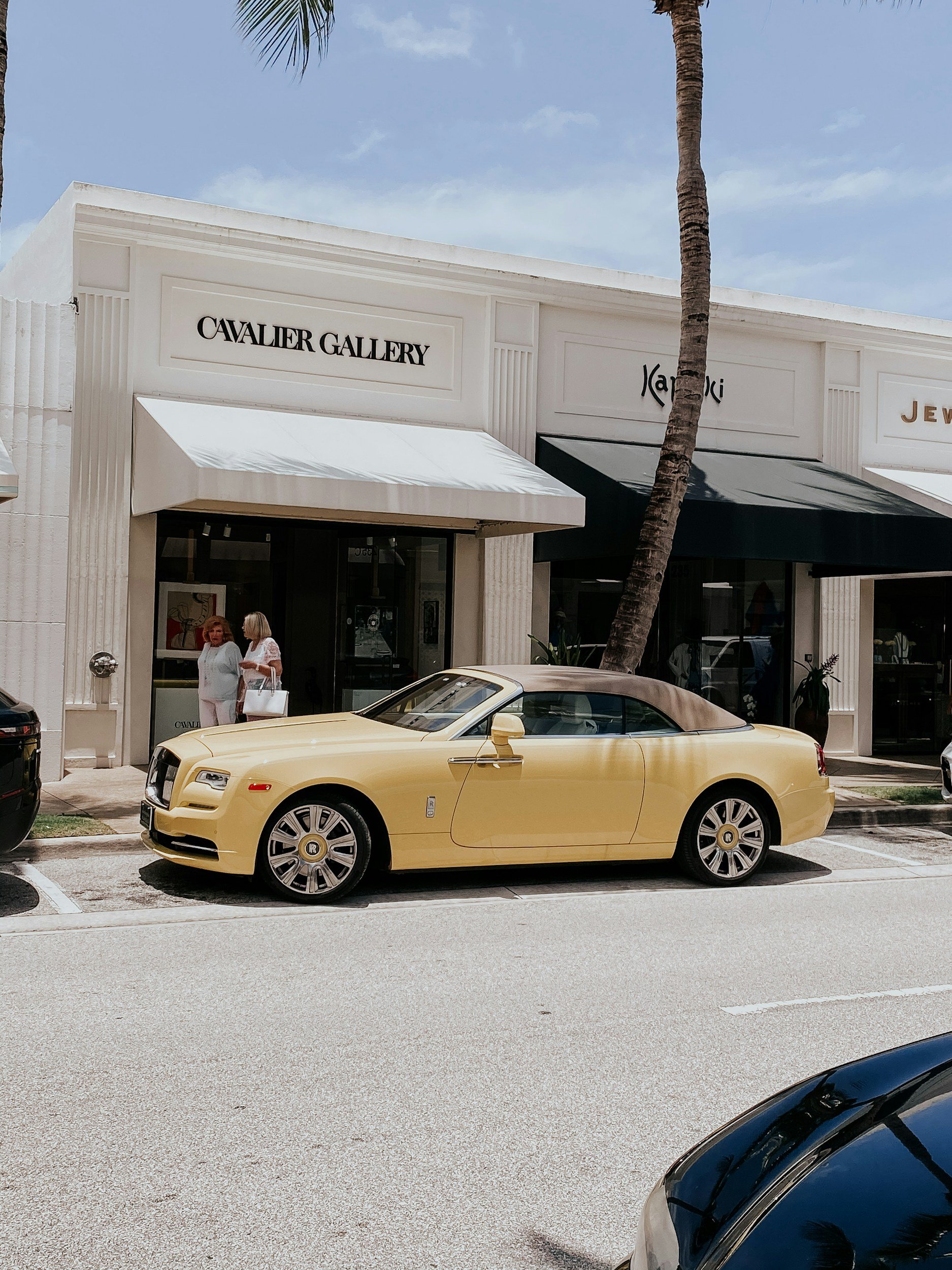 A Palm Beach storefront with signs for Cavalier Gallery, Karen Ki, and jewelry shops, with a yellow Rolls Royce parked in front, palm trees, and two women standing and talking near the entrance.