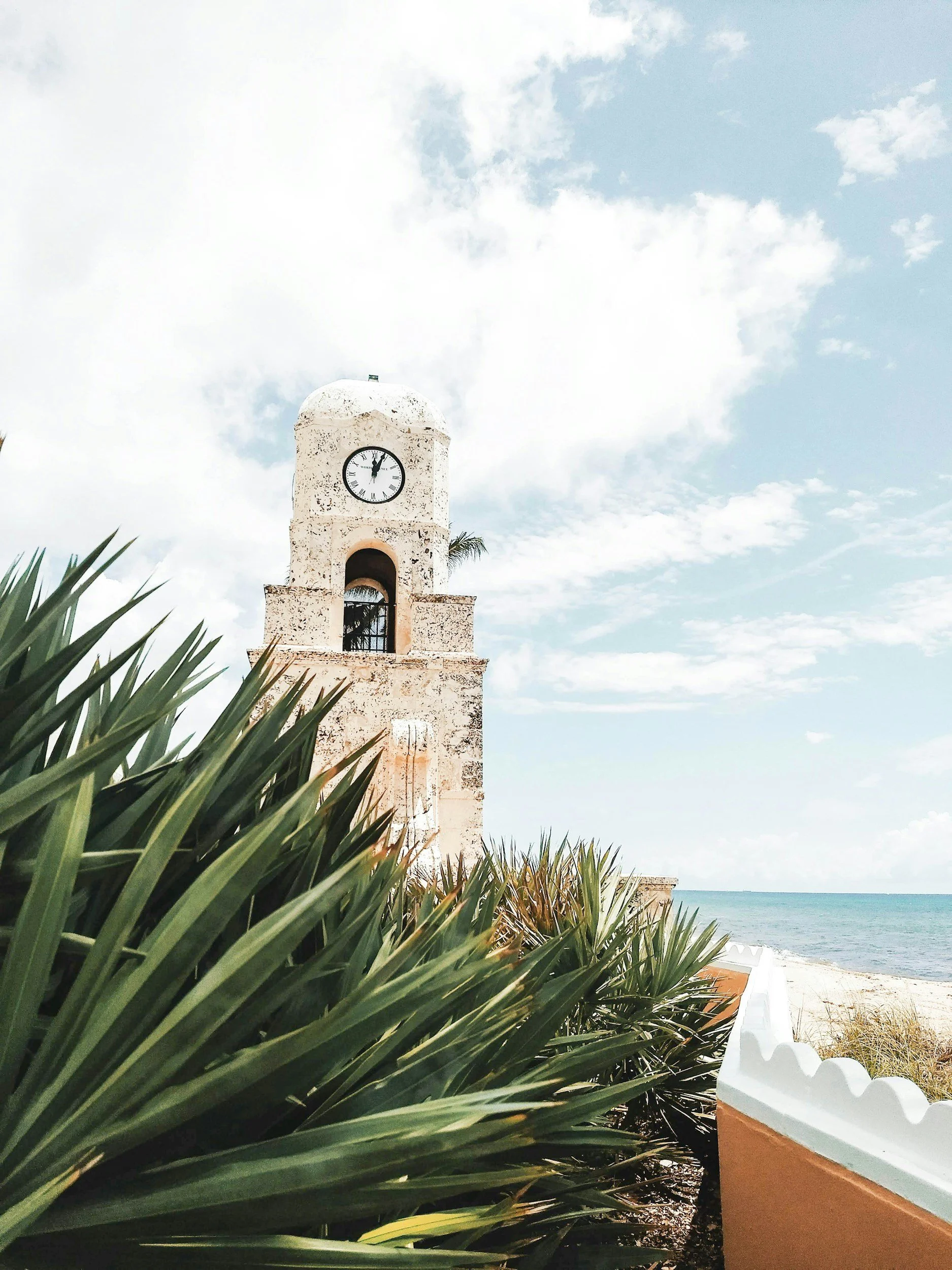 Palm Beach beachfront scene with white clock tower, plants in the foreground, sandy pathway, ocean in the background, partly cloudy sky.