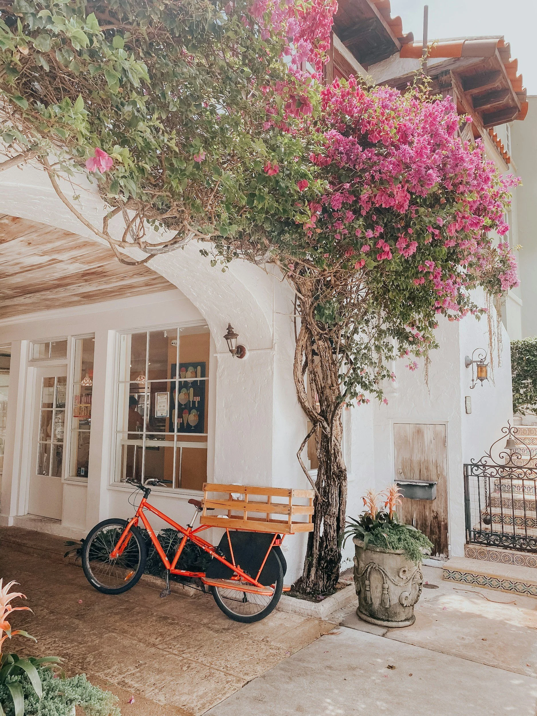 A orange bicycle with a wooden cart attached to the back, parked in front of a white building with a large window and a door. A flowering tree with pink blossoms is growing beside the bicycle, and there are potted plants nearby. The building has a tiled roof and decorative lighting fixtures.