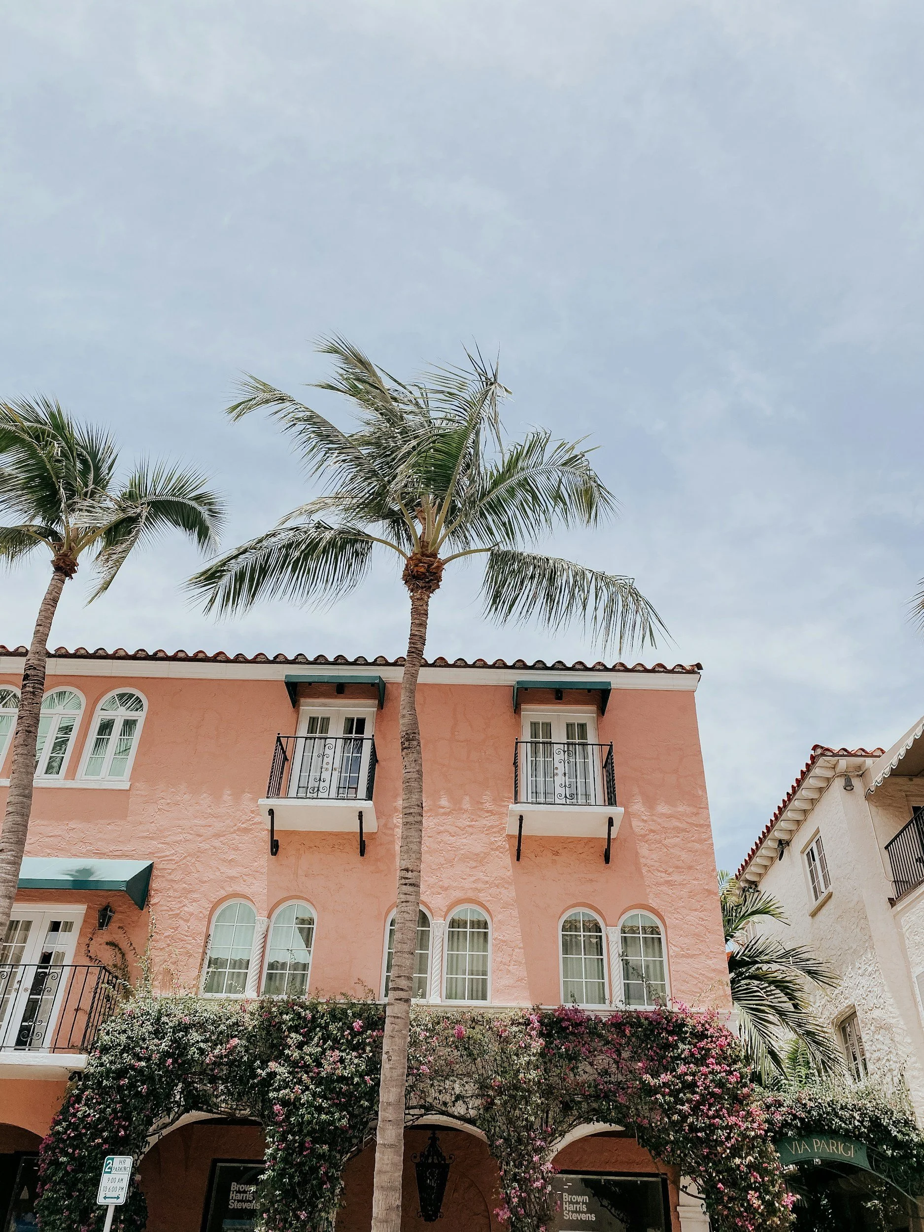 A pink Palm Beach building with arched windows and balconies, two tall palm trees in front, and flowering bushes at the base, under a cloudy sky.