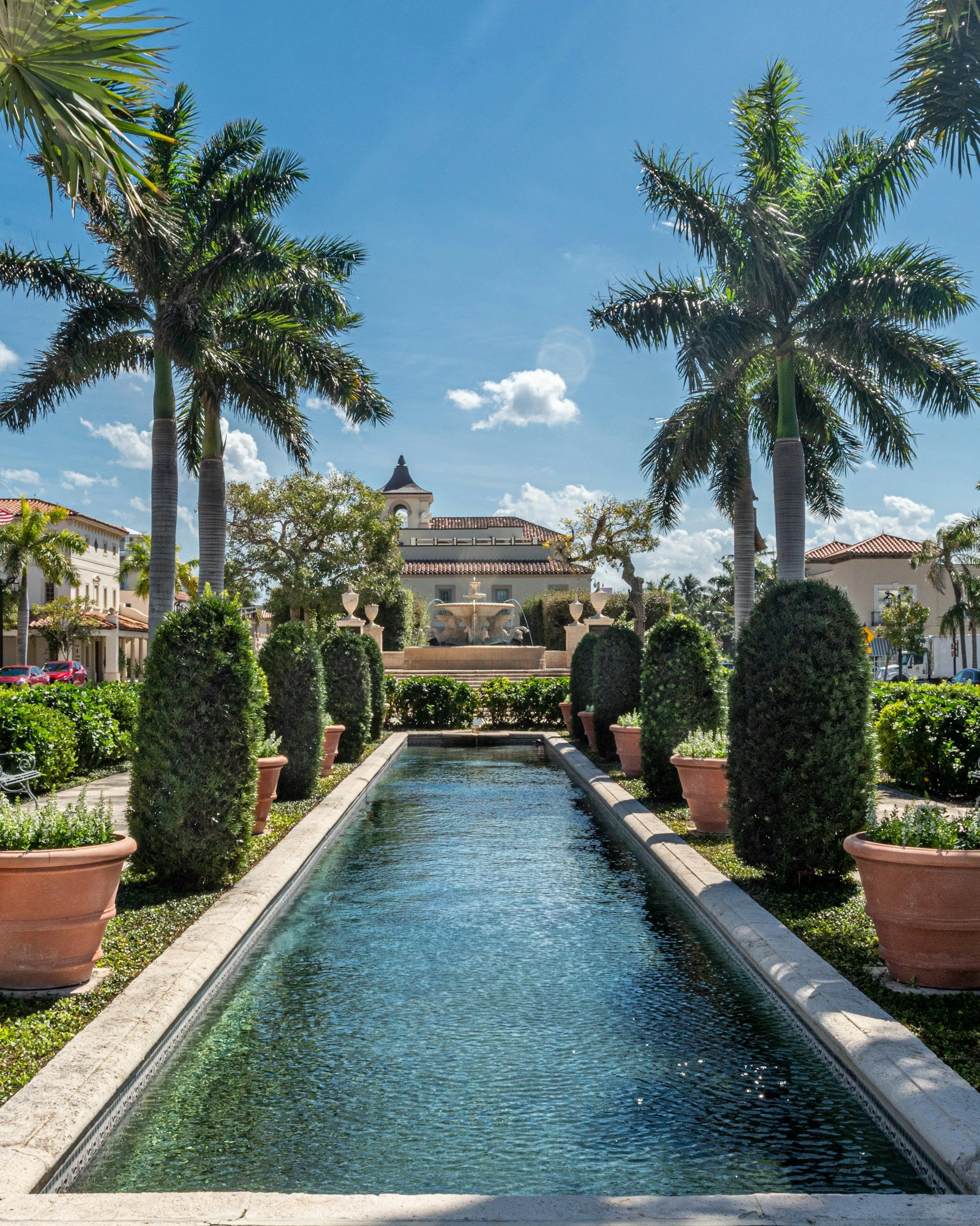 A view of Palm Beach building showing a rectangular water feature with a fountain at the center, surrounded by potted plants and tall palm trees against a bright blue sky.