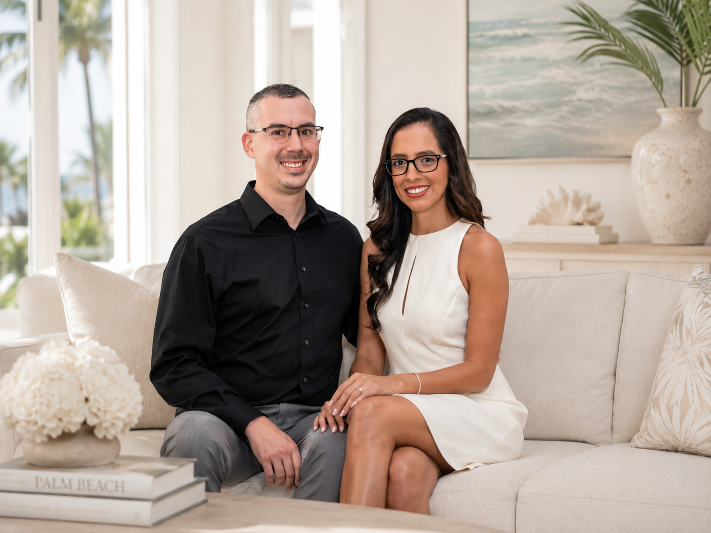 A smiling man and woman sitting together on a beige couch in a bright, modern living room with ocean view outside the window. The man has short hair, glasses, a black shirt, and gray pants. The woman has long dark hair, glasses, a white sleeveless dress, and a ring on her finger.