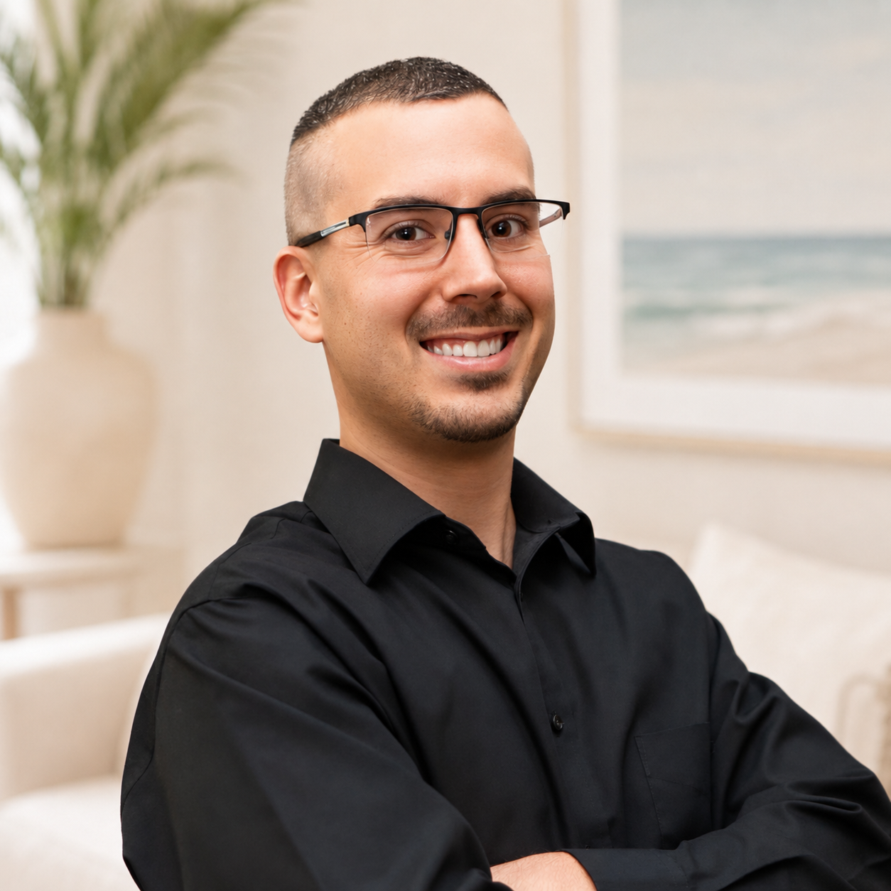 A smiling man (Michael) with short dark hair, glasses, and a black shirt posing with arms crossed in a bright room with a beach painting and a large potted plant in the background.