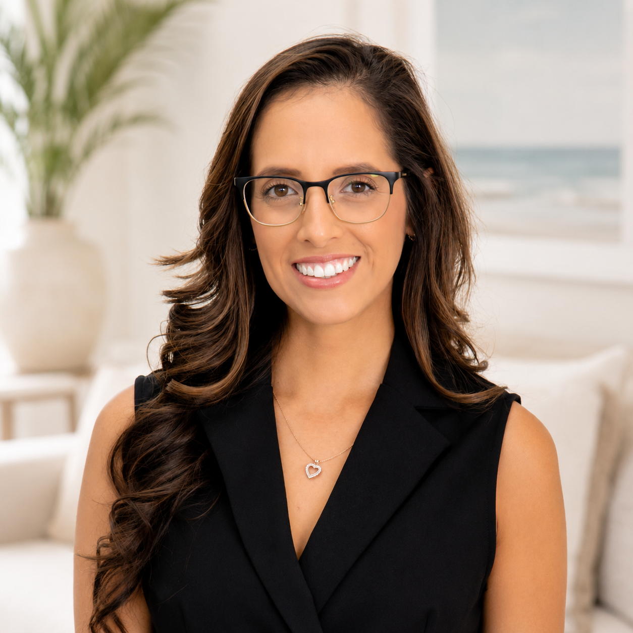 A woman (Pamela) with long brown hair, glasses, and a black sleeveless blazer, smiling in a bright room with a beach scene painting and a potted plant in the background.