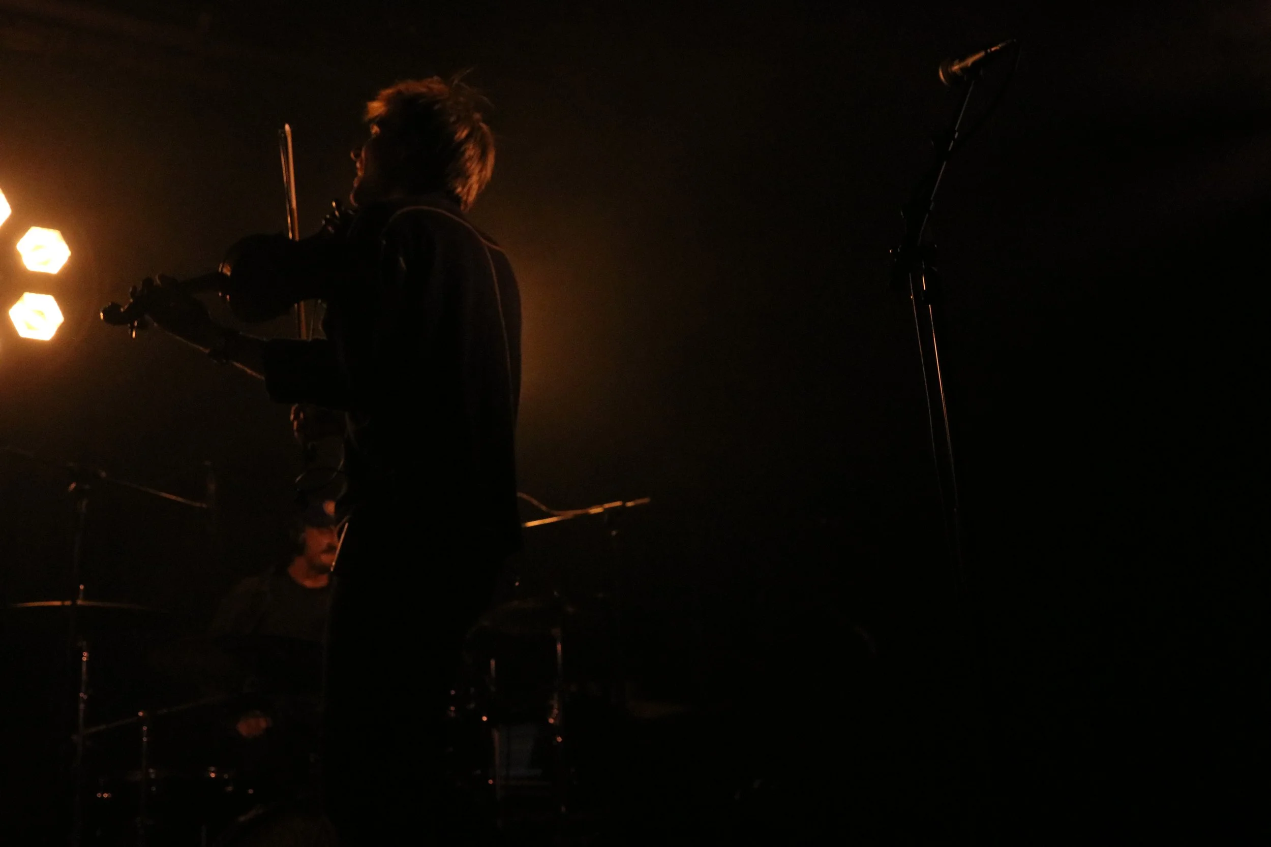 A silhouette of a person playing the violin on stage, with stage lights in the background and a drum set partially visible behind them, with a microphone on a stand nearby.