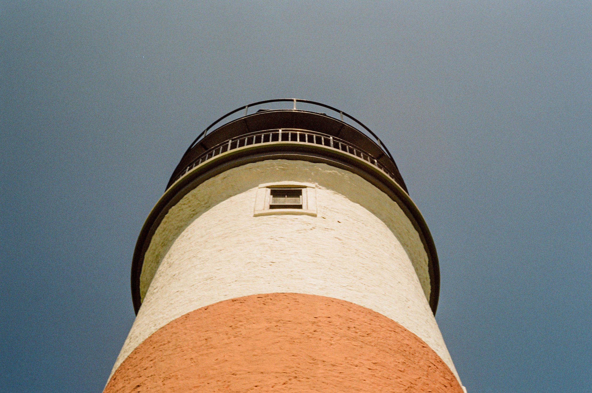 A lighthouse with a white, pink, and greenish exterior against a clear blue sky, viewed from below.