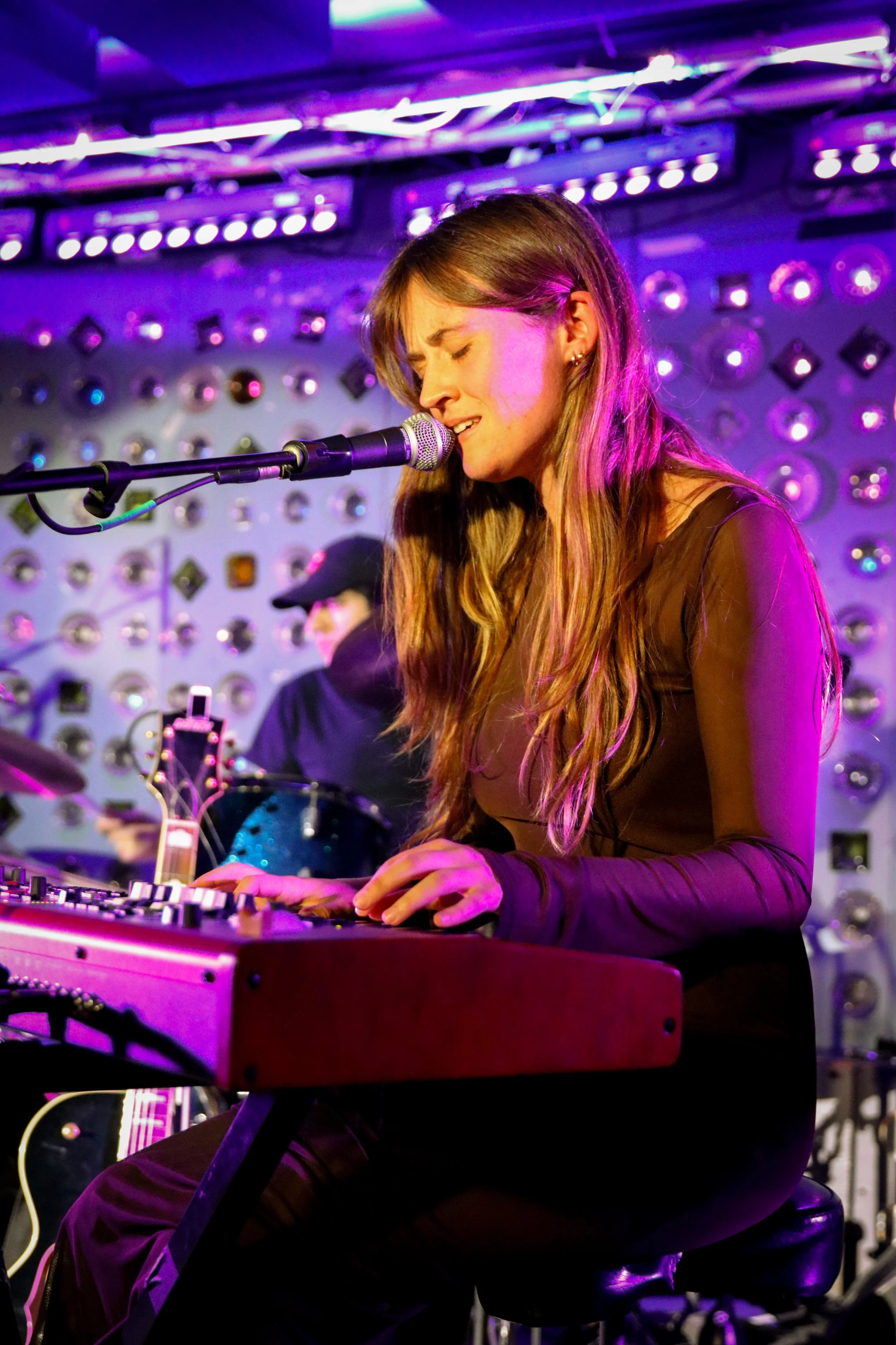 A woman with long wavy hair wearing a sheer black top is playing a keyboard and singing into a microphone. She appears emotional with her eyes closed. There is a person in the background playing a drum, all illuminated by purple stage lighting.