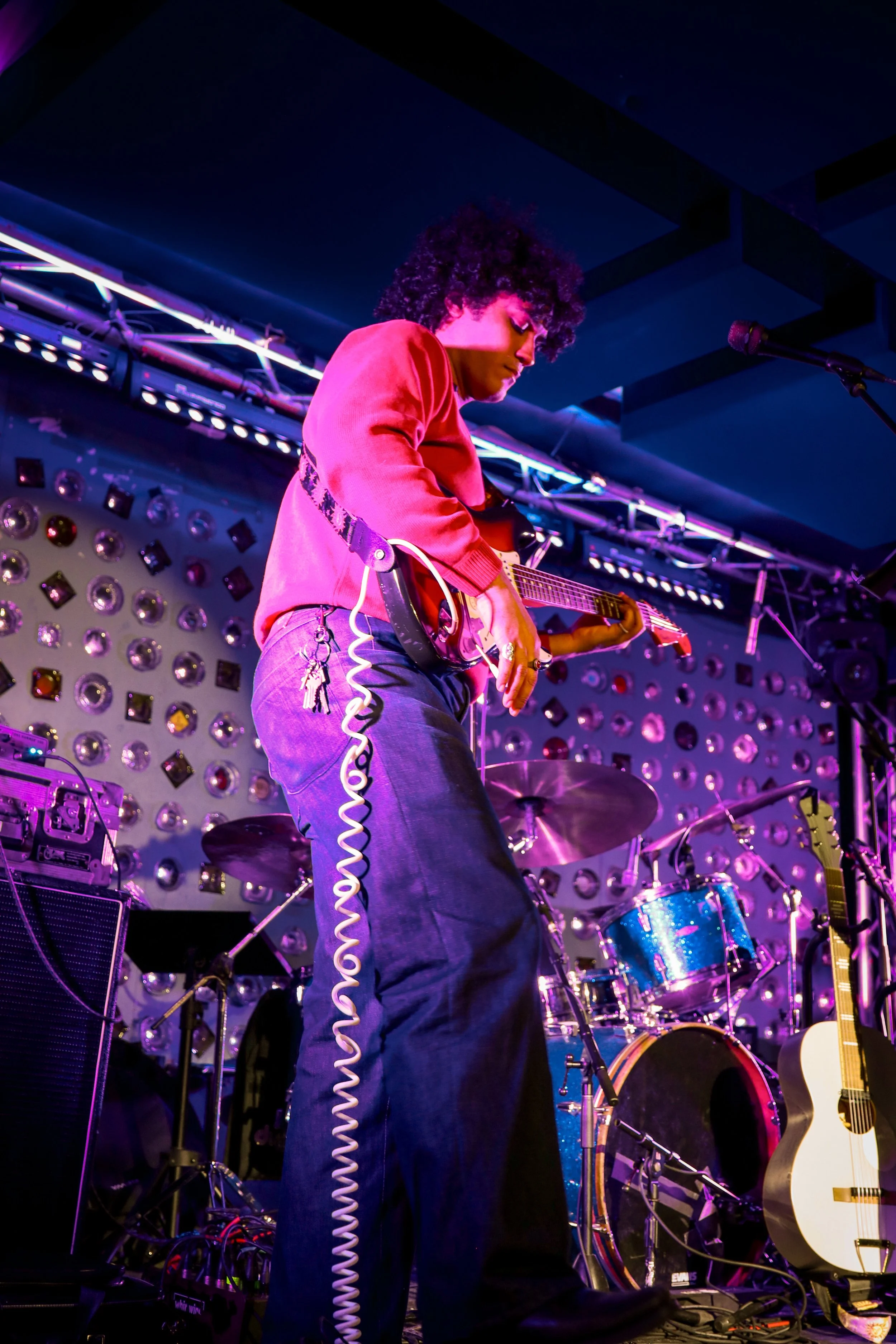 A musician with curly hair wearing a red shirt playing an electric guitar on stage with colorful lighting and musical instruments in the background.