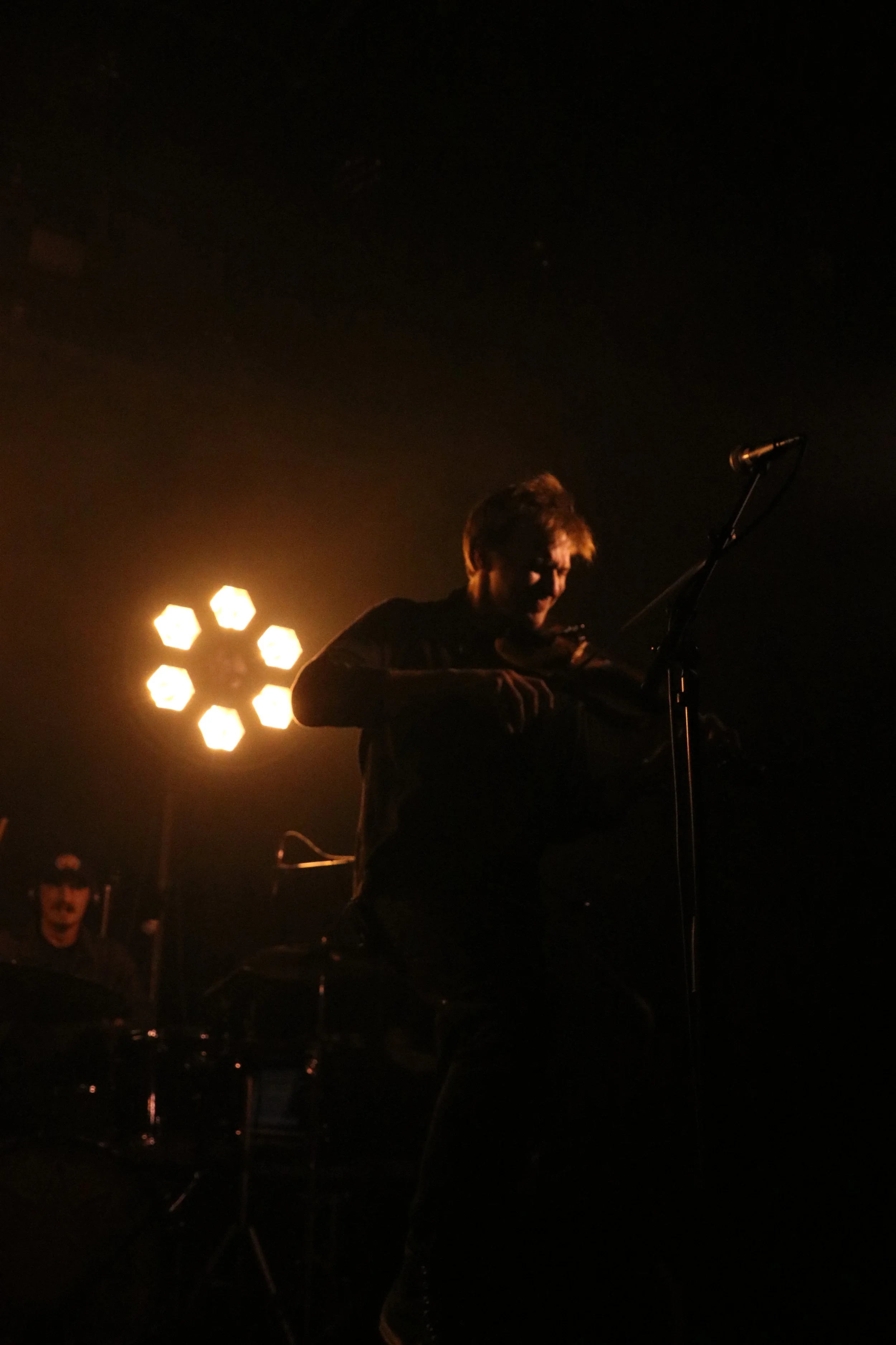 A person playing a violin on stage with a bright light behind them, and another person in the background at a drum set.