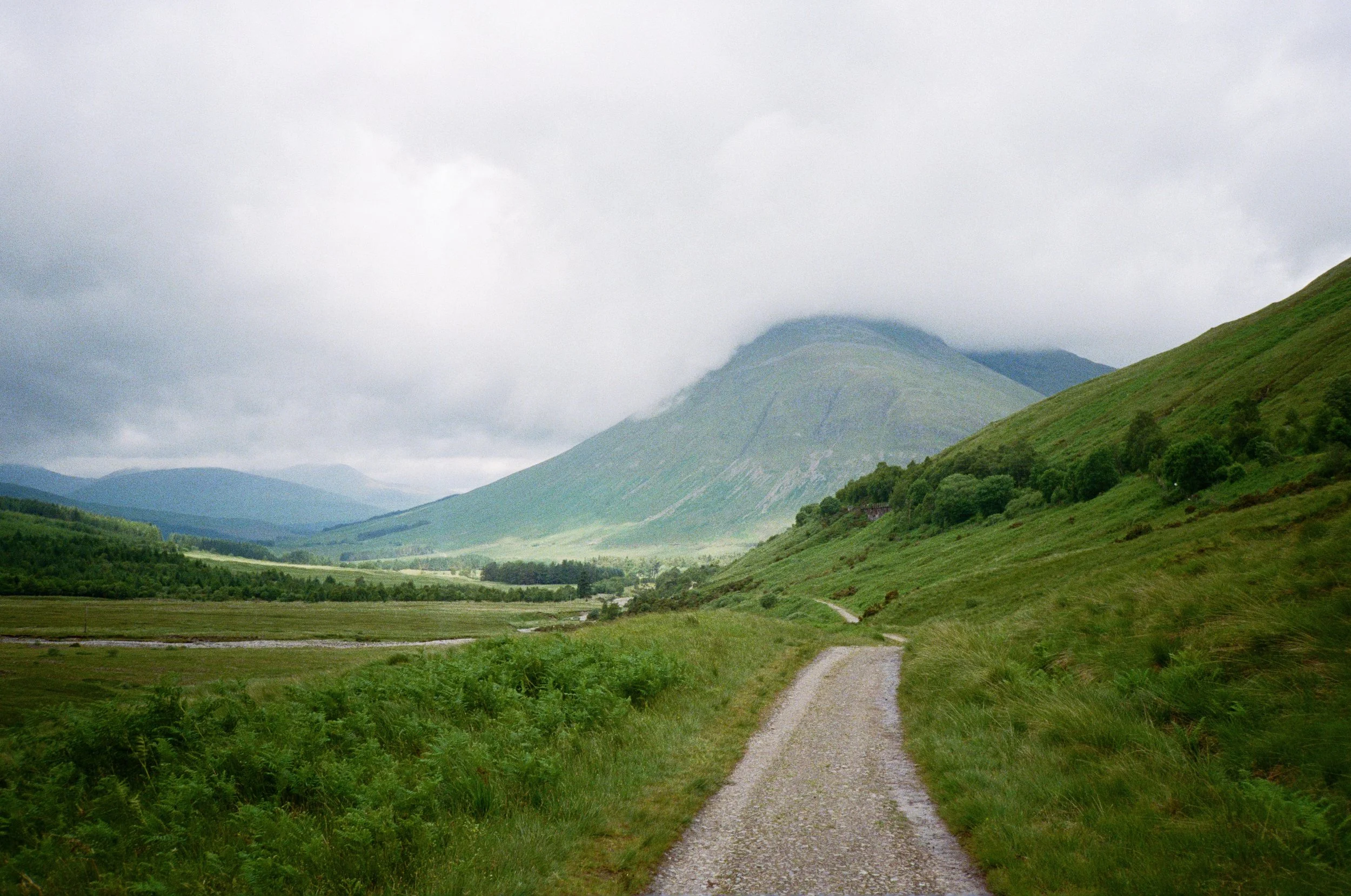 A gravel path winding through lush green fields and hills with a mountain partly covered by clouds in the background under a gray, cloudy sky.