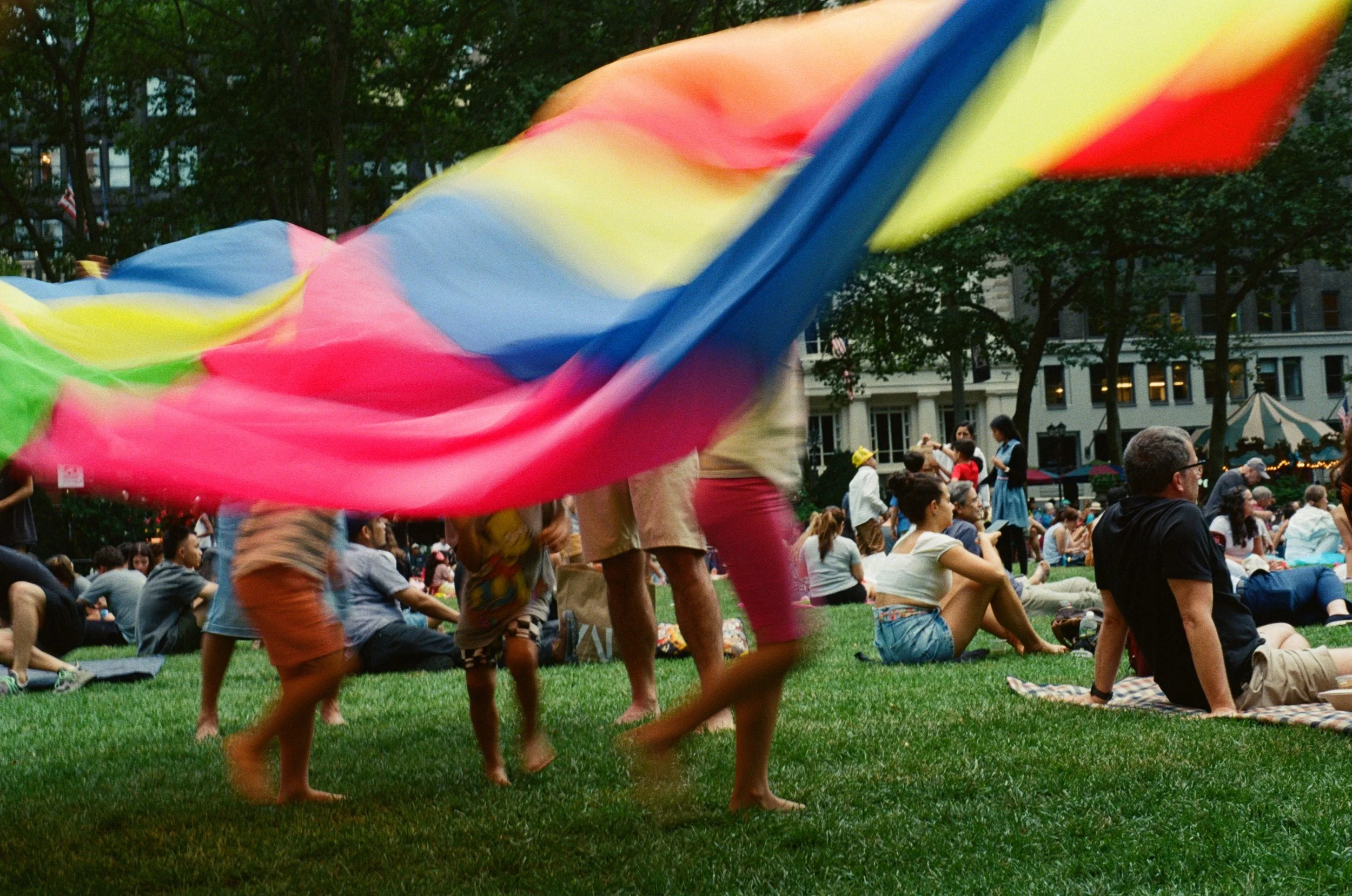 Children playing with a multicolored parachute in a park, surrounded by people sitting on grass and city buildings in the background.