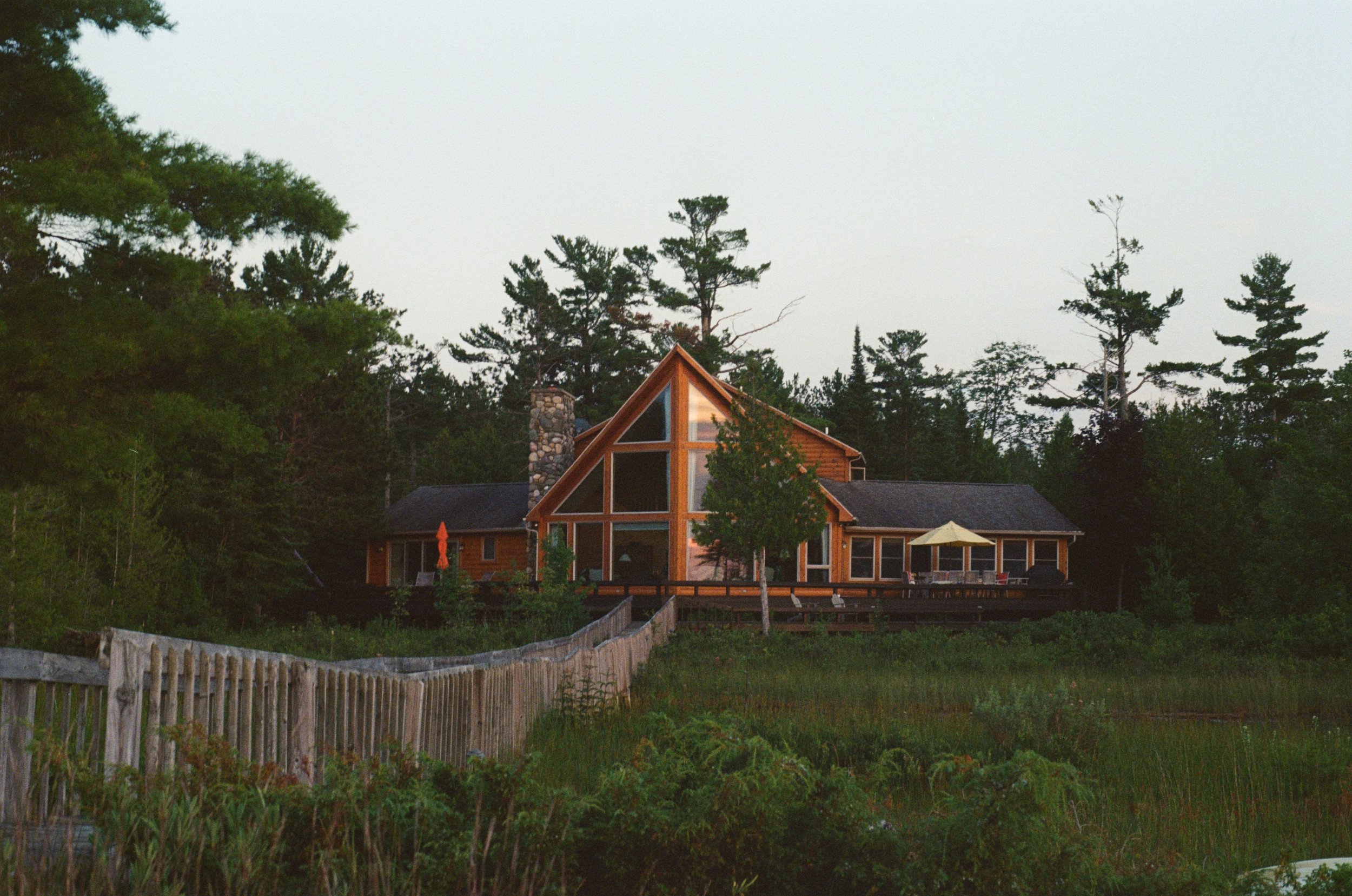 A wooden house with large triangular glass windows, a chimney, and a deck with a yellow umbrella, surrounded by trees and greenery.