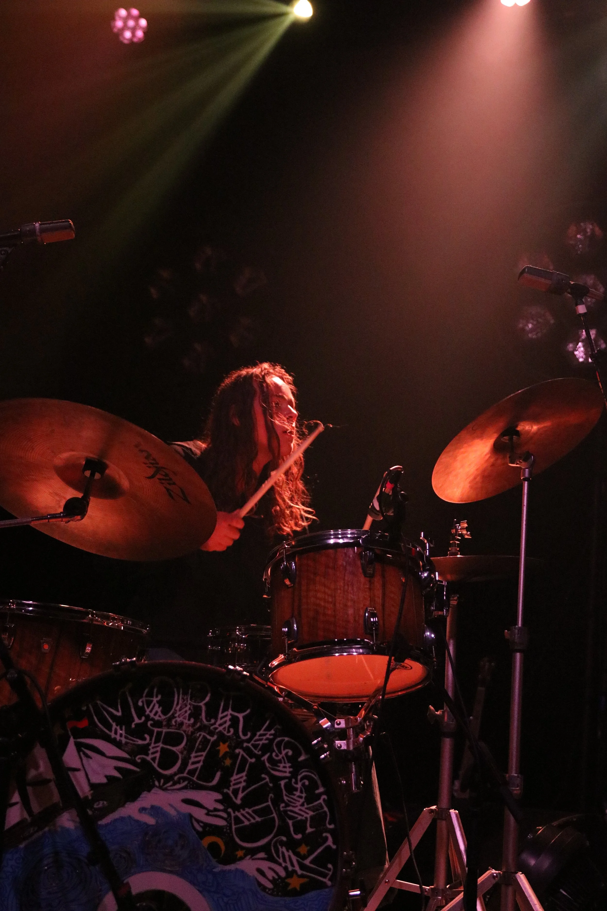 A drummer performing on stage with long hair, illuminated by colorful stage lights.