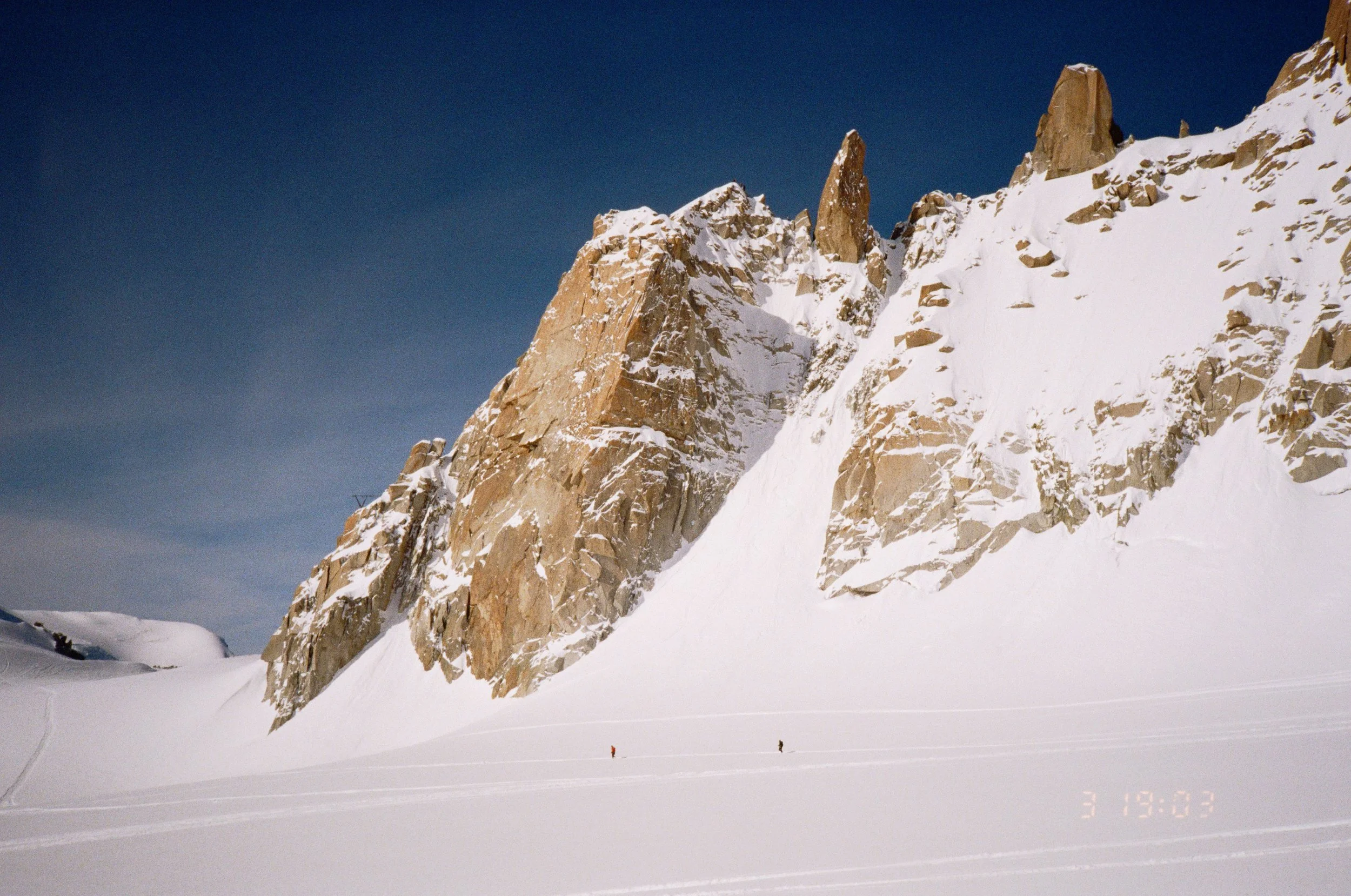 Snow-covered mountain landscape with rugged peaks and two small figures in the snow at the bottom, under a clear blue sky.