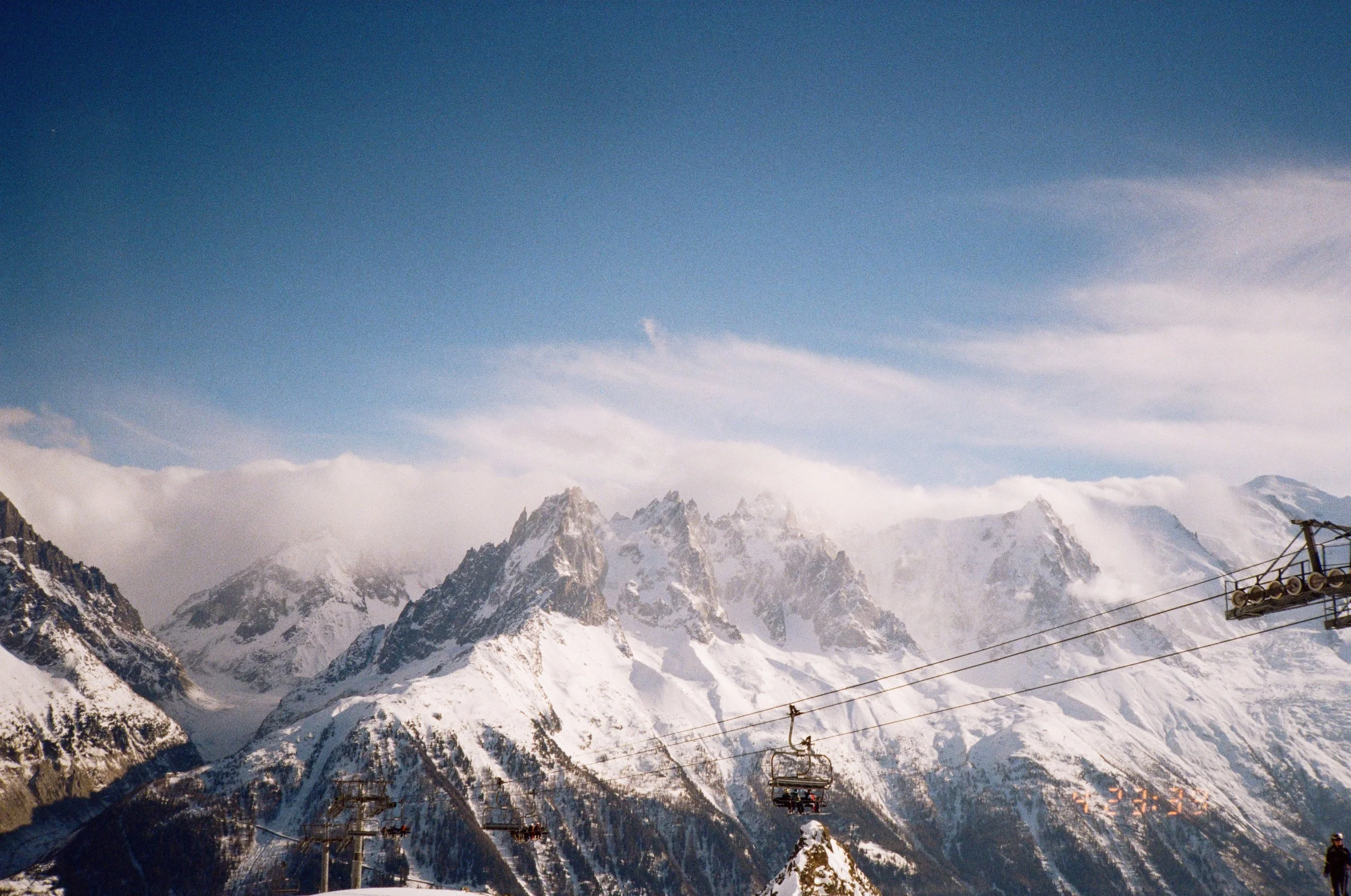 Snow-covered mountains with a ski lift and cables in the foreground under a blue sky with few clouds.