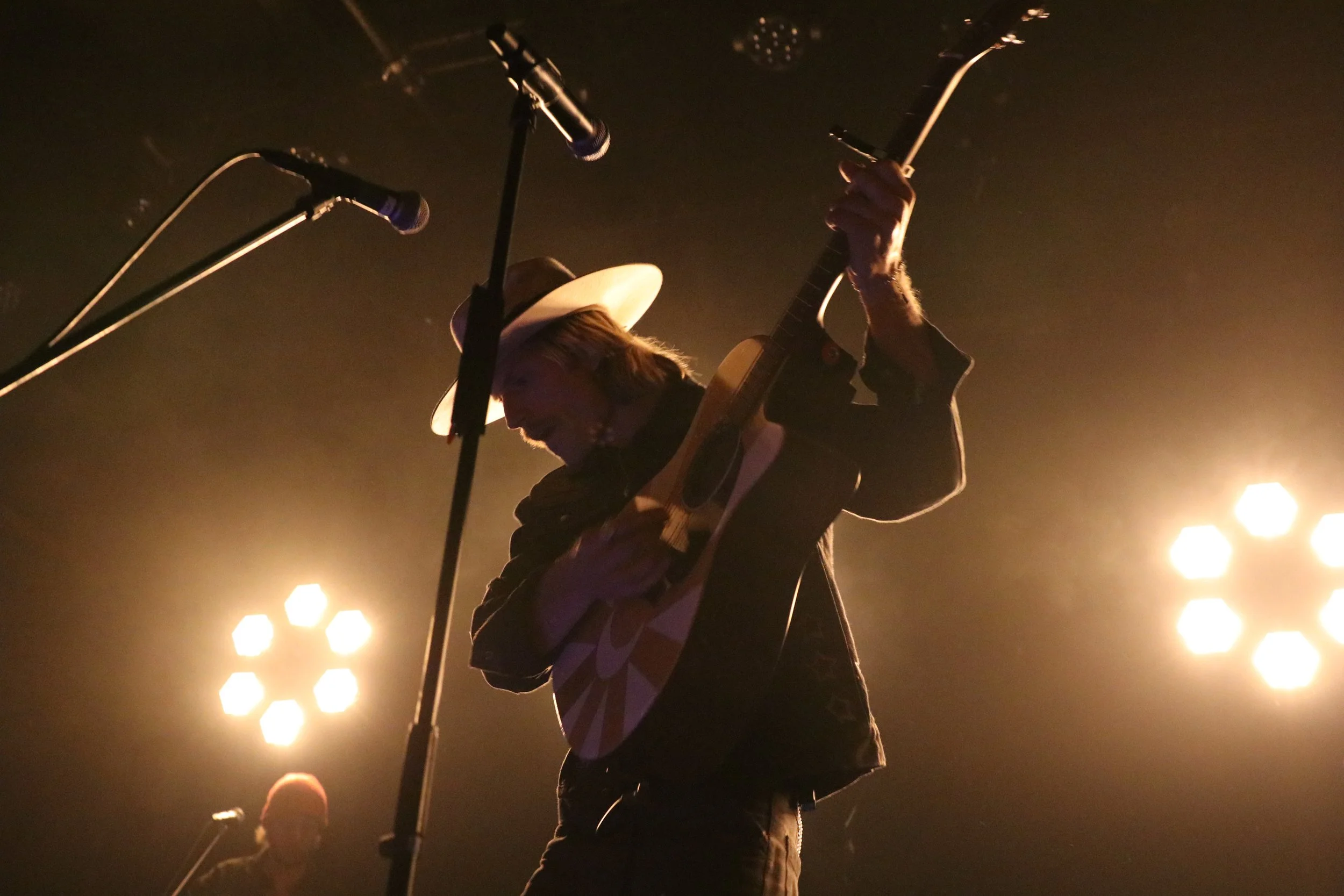 A musician wearing a wide-brimmed hat playing an acoustic guitar on stage, surrounded by bright stage lights.
