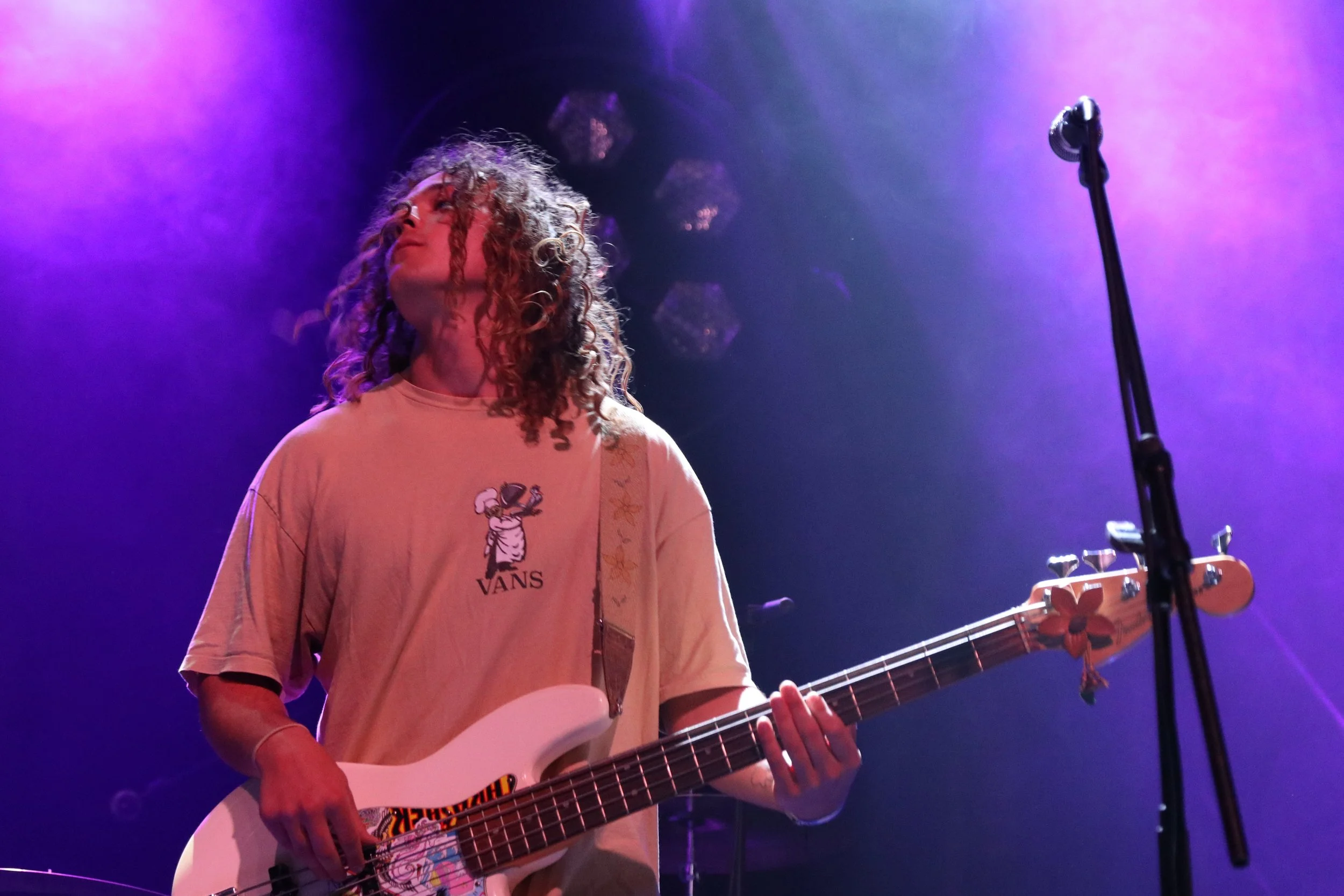 A young woman with curly hair playing an electric bass guitar on stage under purple lights.