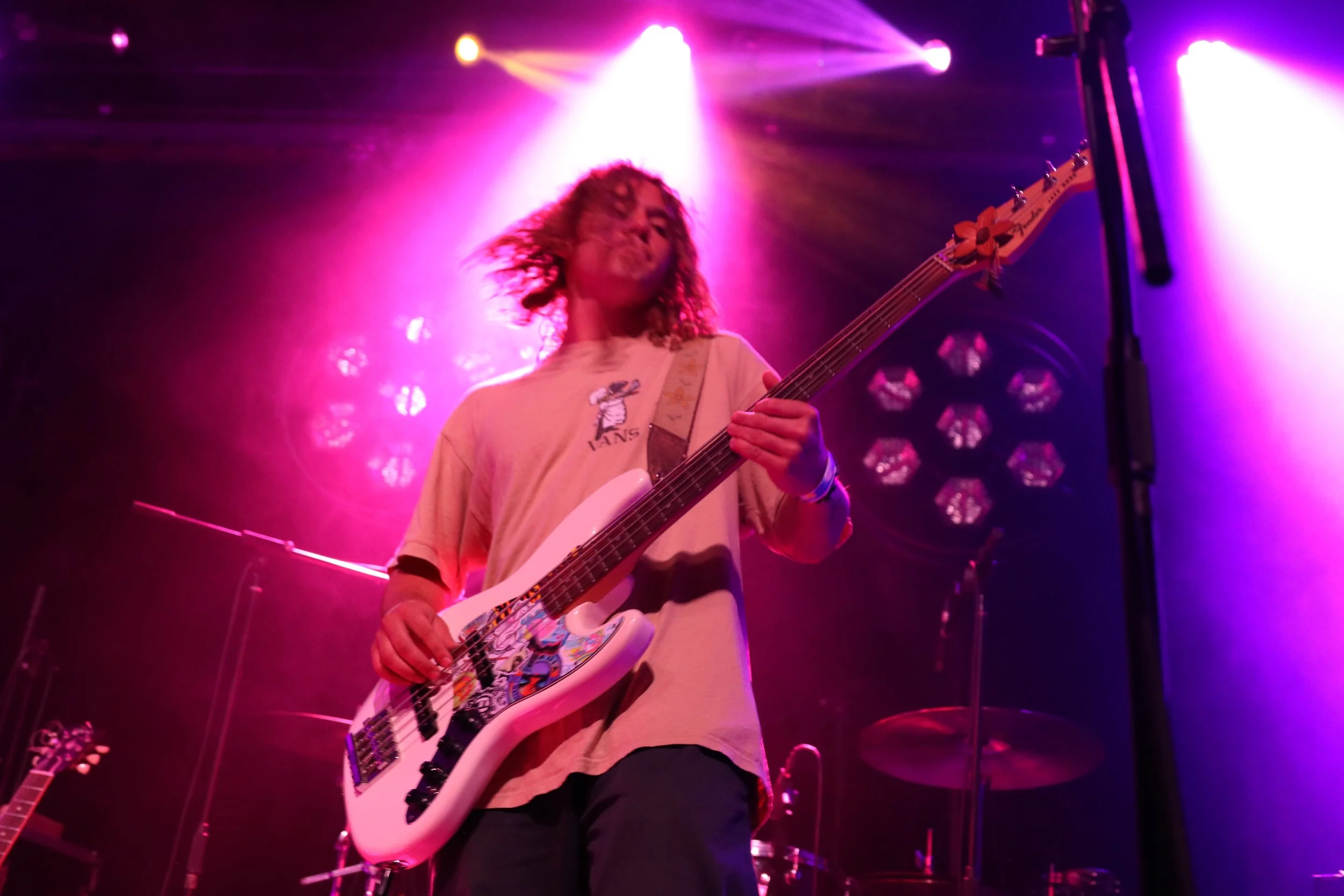 A person playing electric guitar on stage under colorful pink and purple lighting with a drum set in the background.