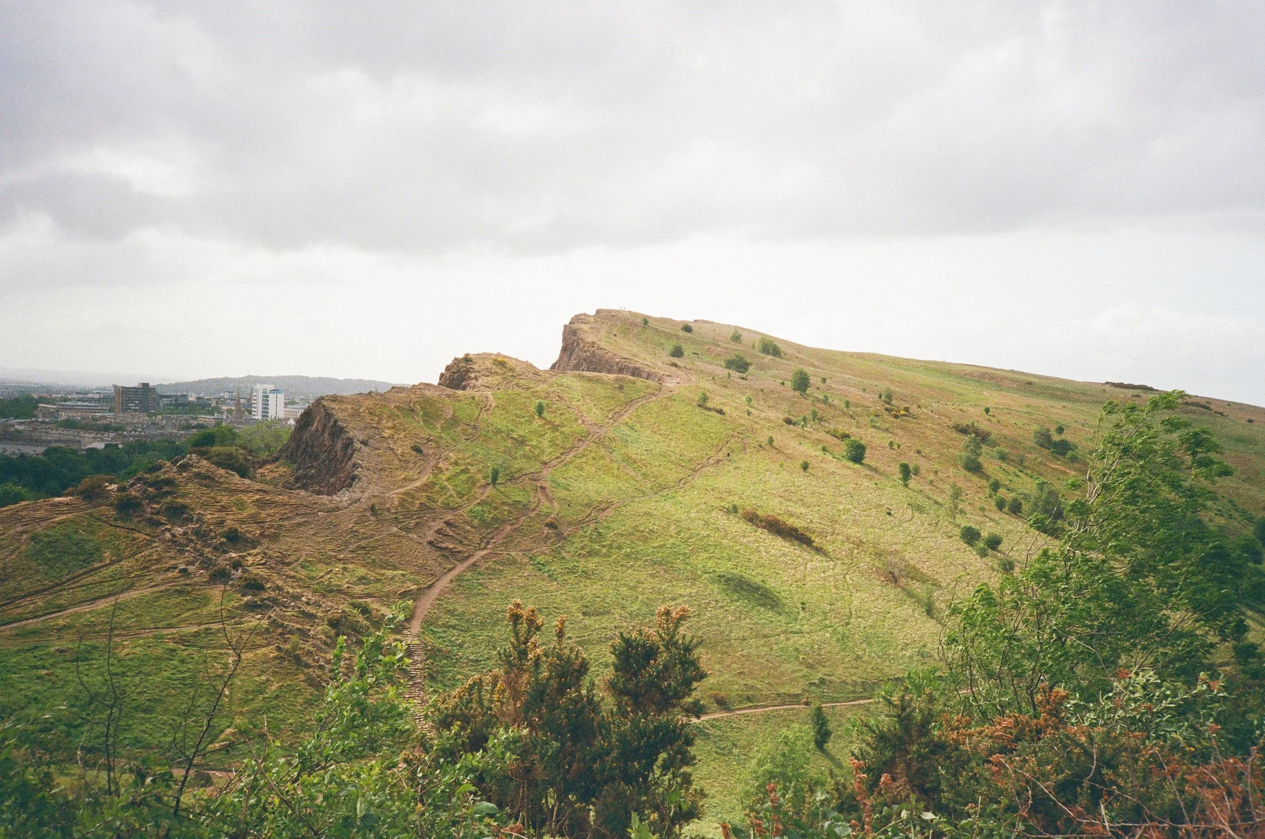 A photo of the volcanic hill Arthur's Seat in Edinburgh, Scotland, with hiking trails and sparse vegetation under a cloudy sky.