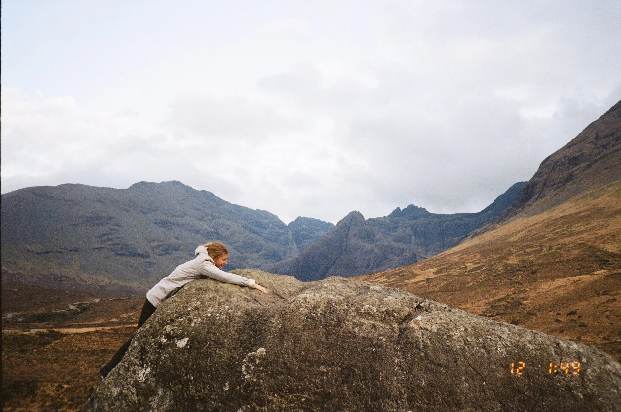 A woman in a grey jacket leaning on a large rock in a mountainous landscape with cloudy skies, brown and green hills, and distant mountains.