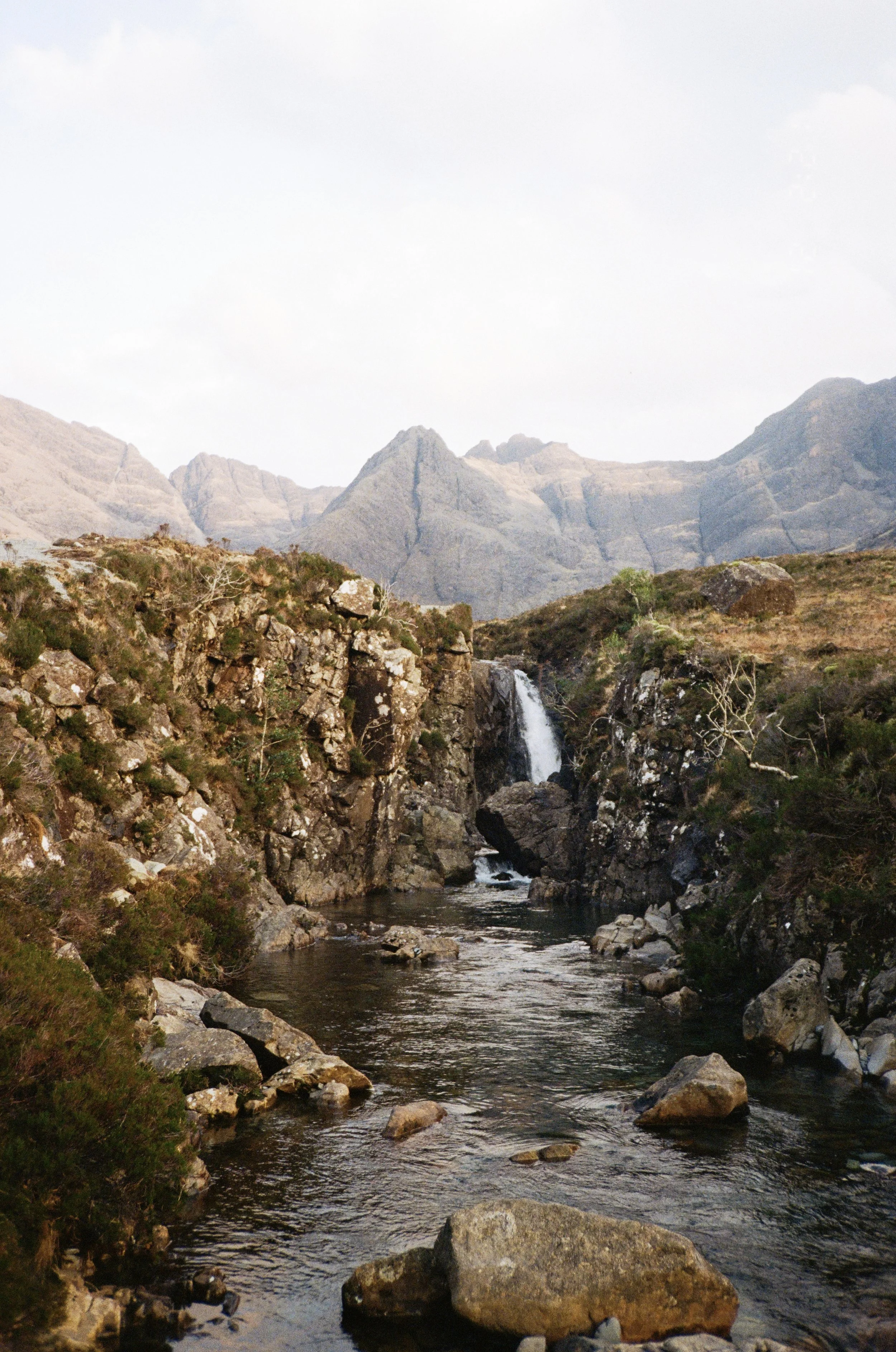 A river flowing through rocky terrain with a small waterfall and mountain range in the background under a cloudy sky.