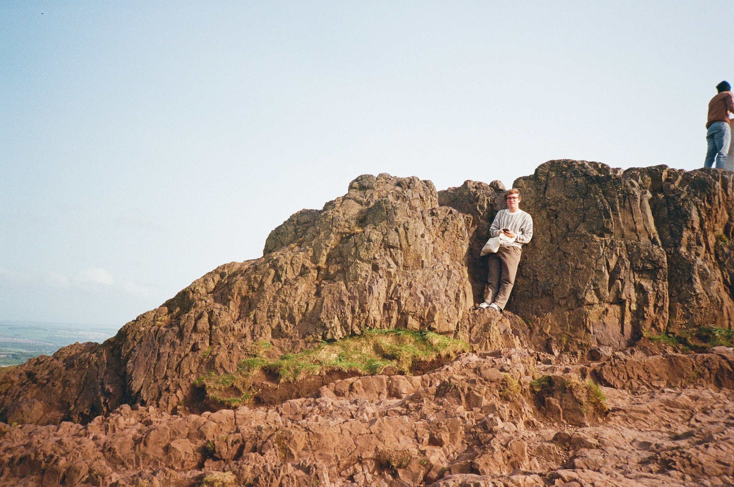 Person sitting on a large rocky hillside, looking at the camera, with two other people standing on the rocks nearby, under a clear sky.