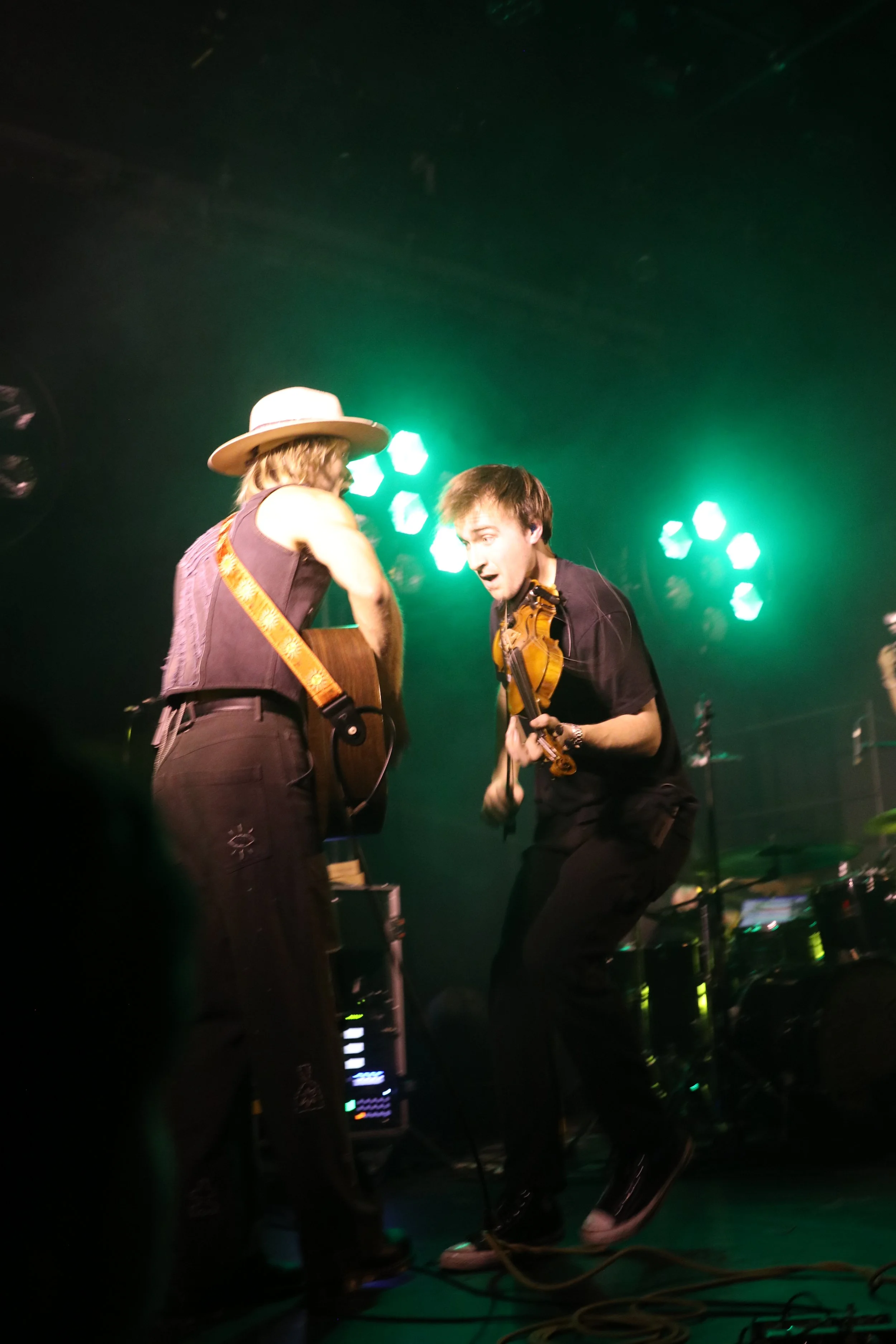 Two musicians performing on stage, one playing an acoustic guitar and the other playing a violin, with green stage lights in the background.