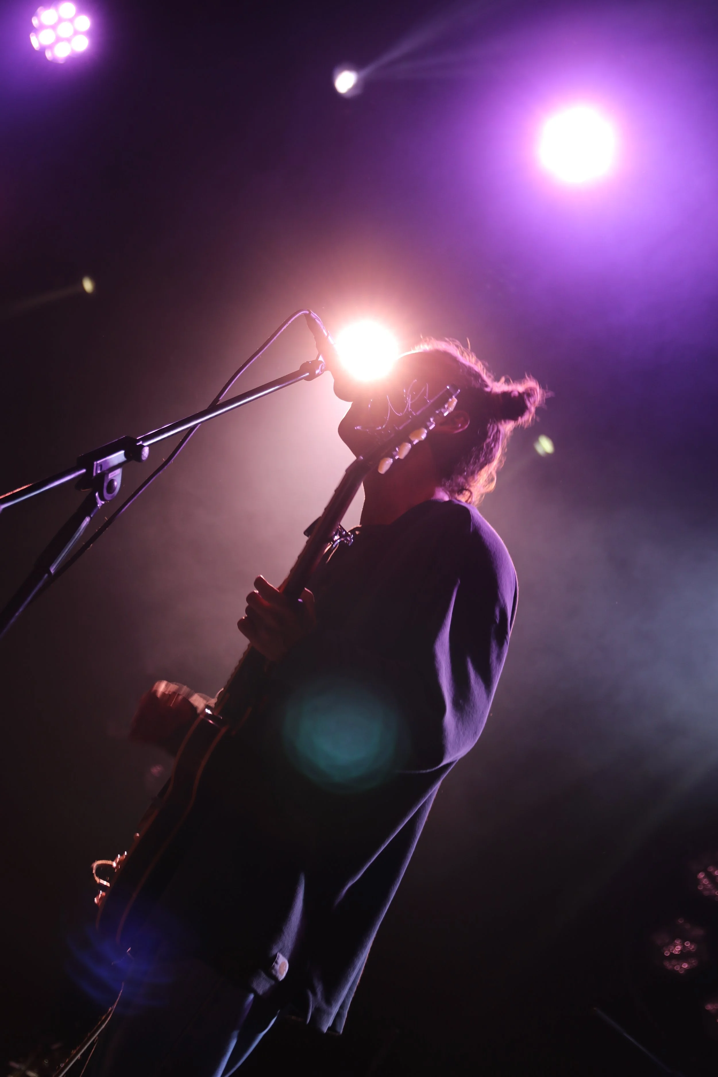 A musician playing guitar on stage with bright purple and white stage lights creating a silhouette effect.