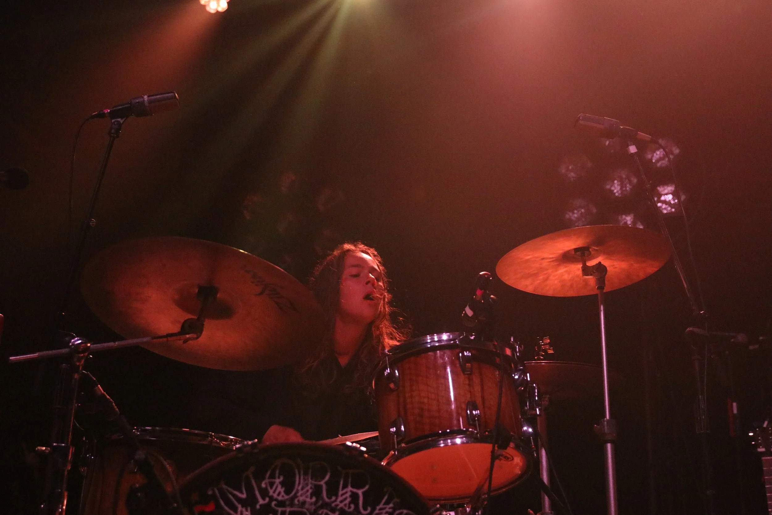 A female drummer playing a drum set on stage with two guitars and microphones. Stage lighting with warm colors is visible.