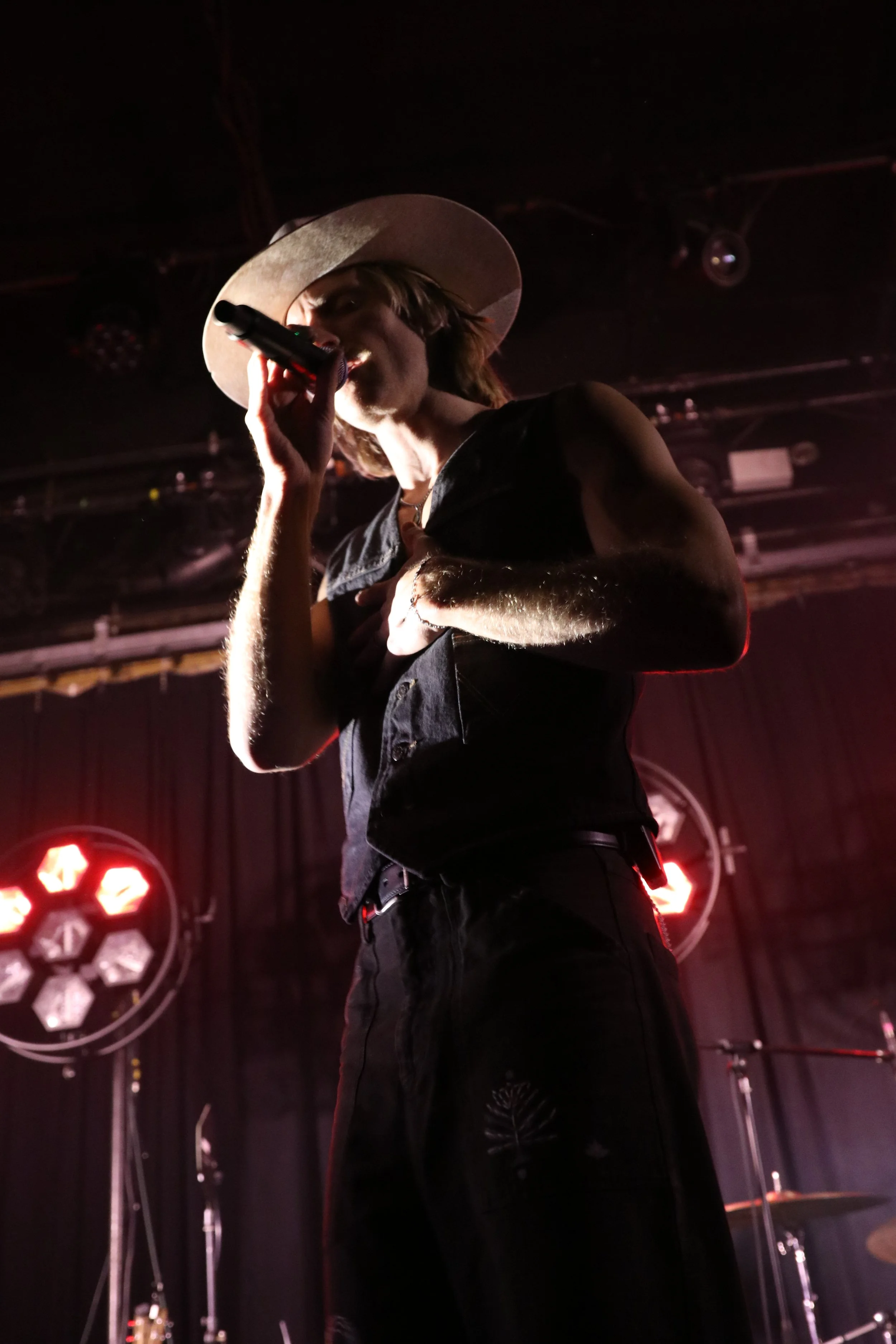 A singer wearing a large hat and sleeveless black outfit performing on stage with red stage lights in the background.