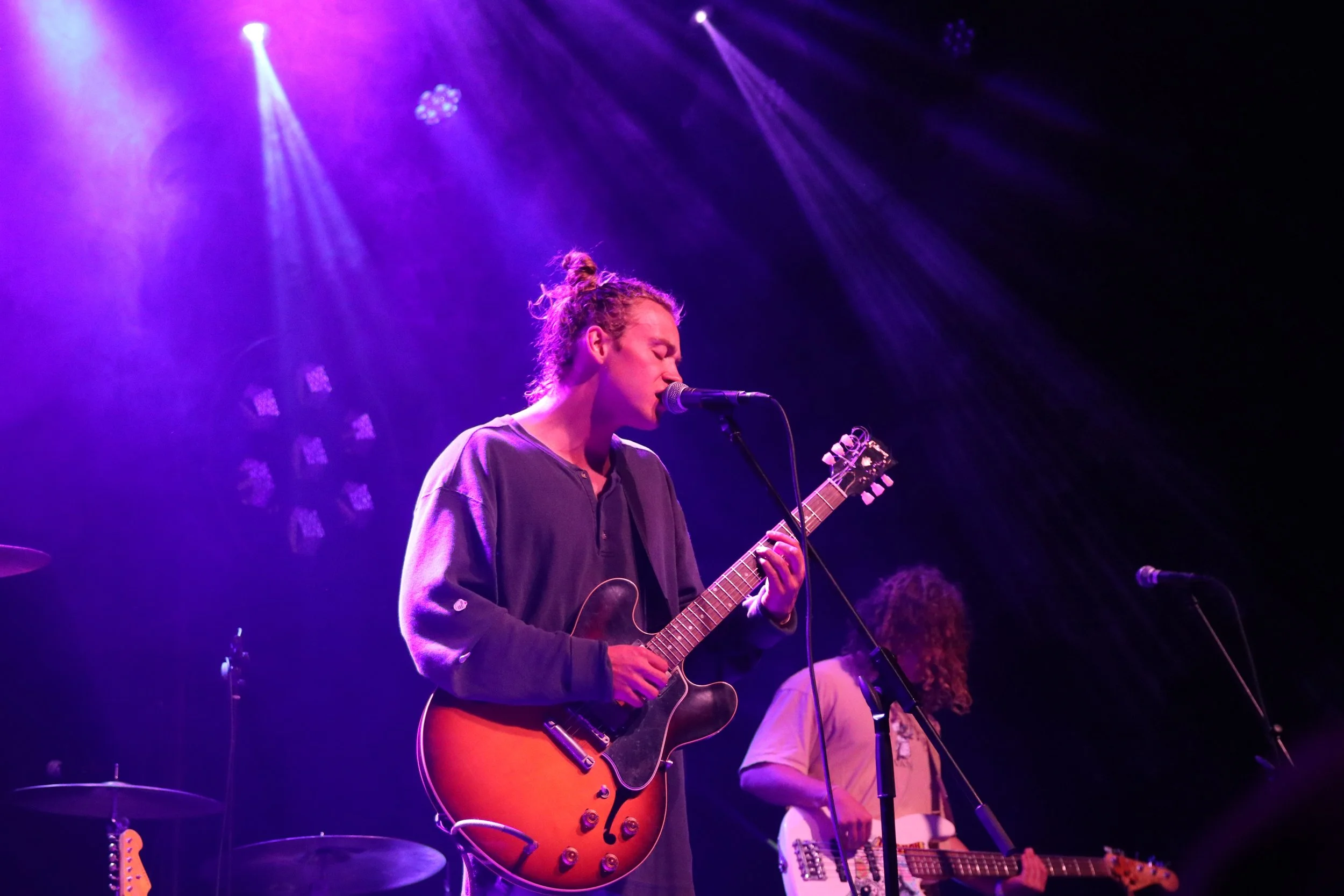 Musician with curly hair playing an electric guitar and singing into a microphone on stage with purple lighting.