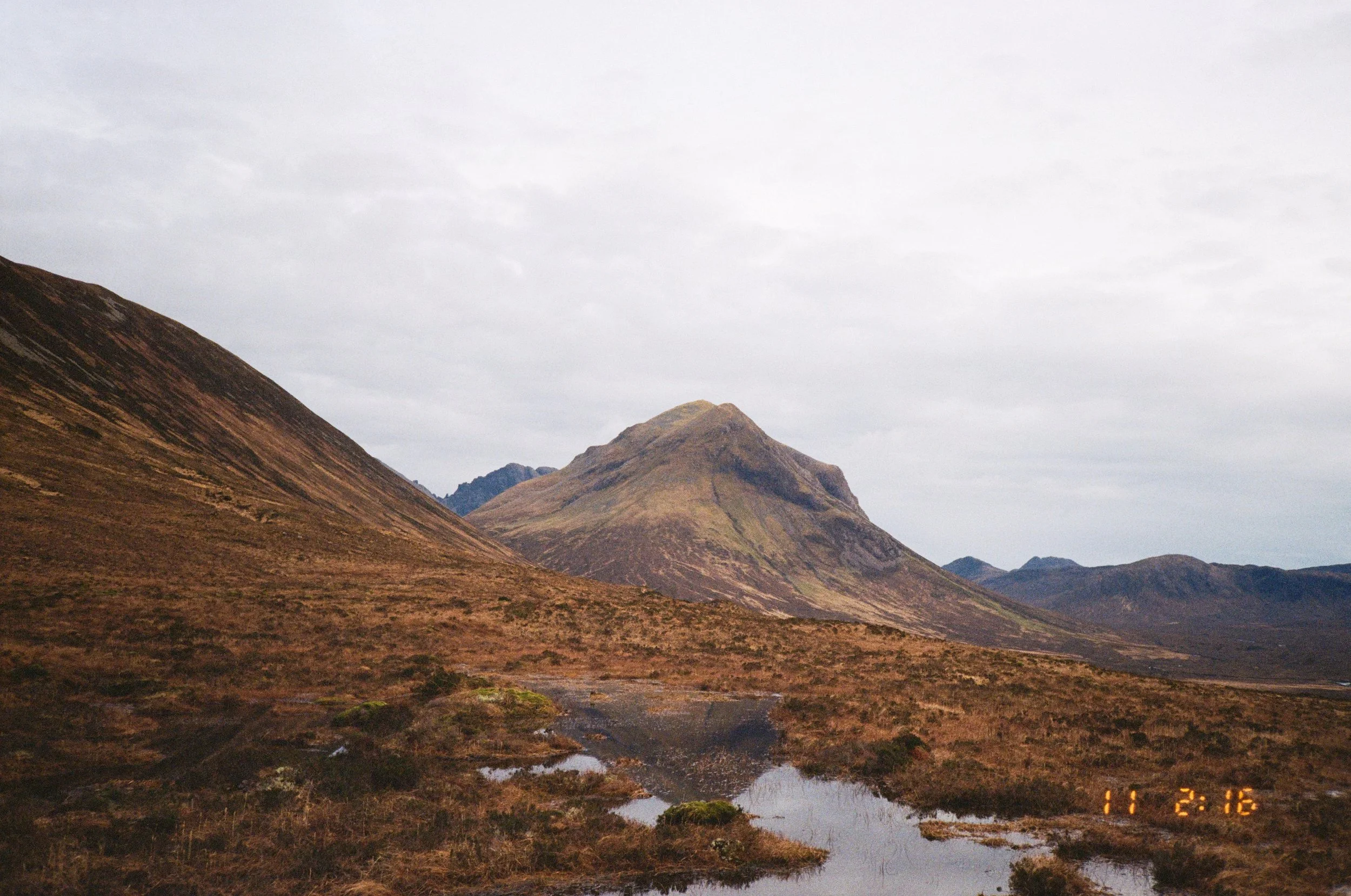 A mountainous landscape with brown and green-covered hills, a small water stream, and a cloudy sky.