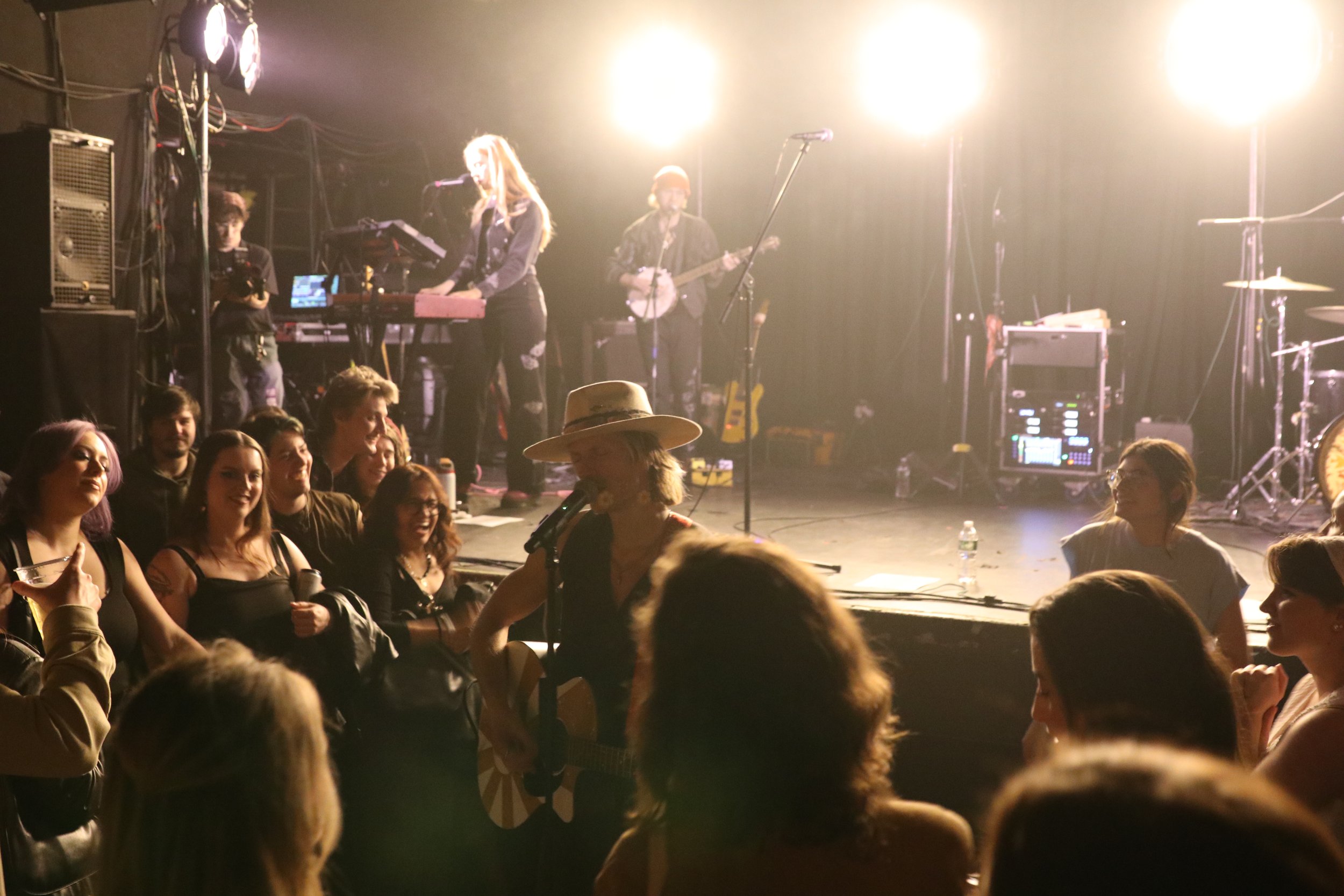 Musicians performing on stage at a concert with a crowd of fans watching, some smiling and holding drinks, and a woman singing with a guitar in the foreground.