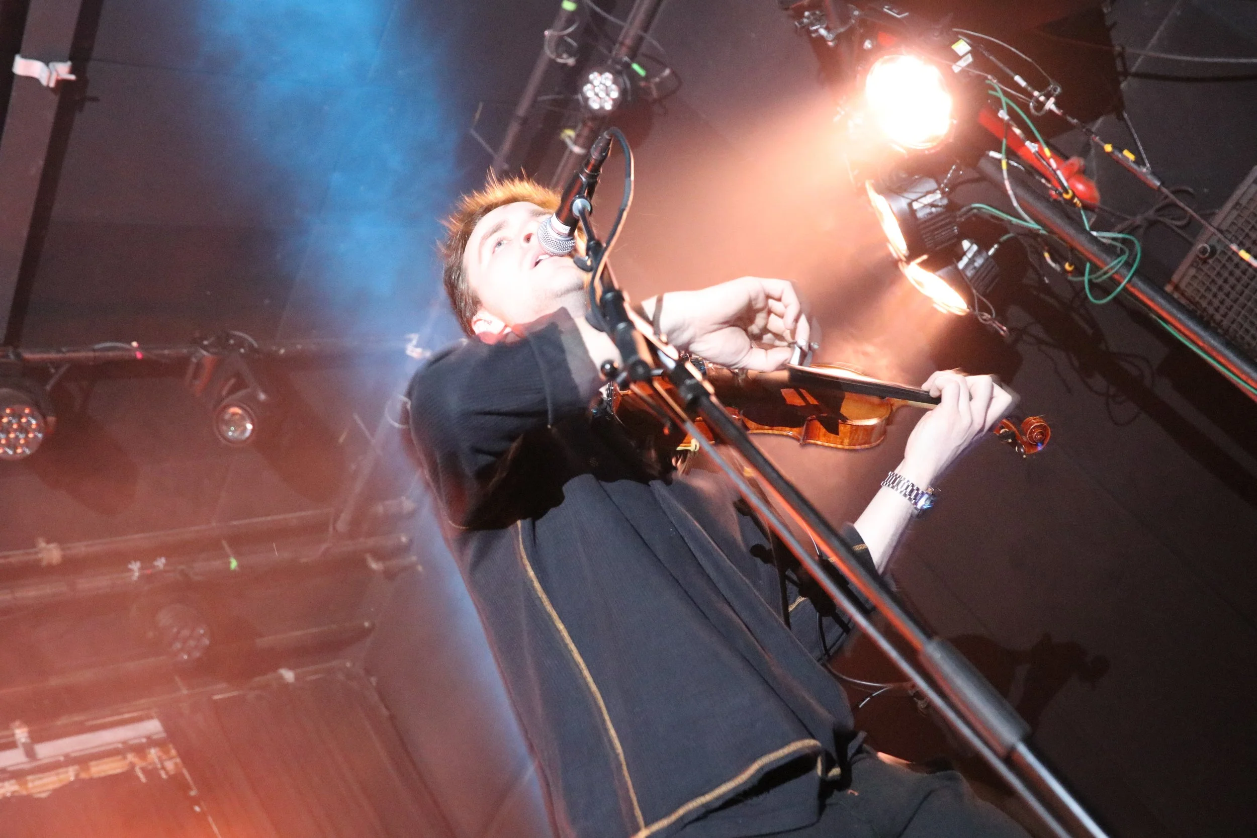 A musician playing a violin on stage with microphone, stage lights, and equipment visible.