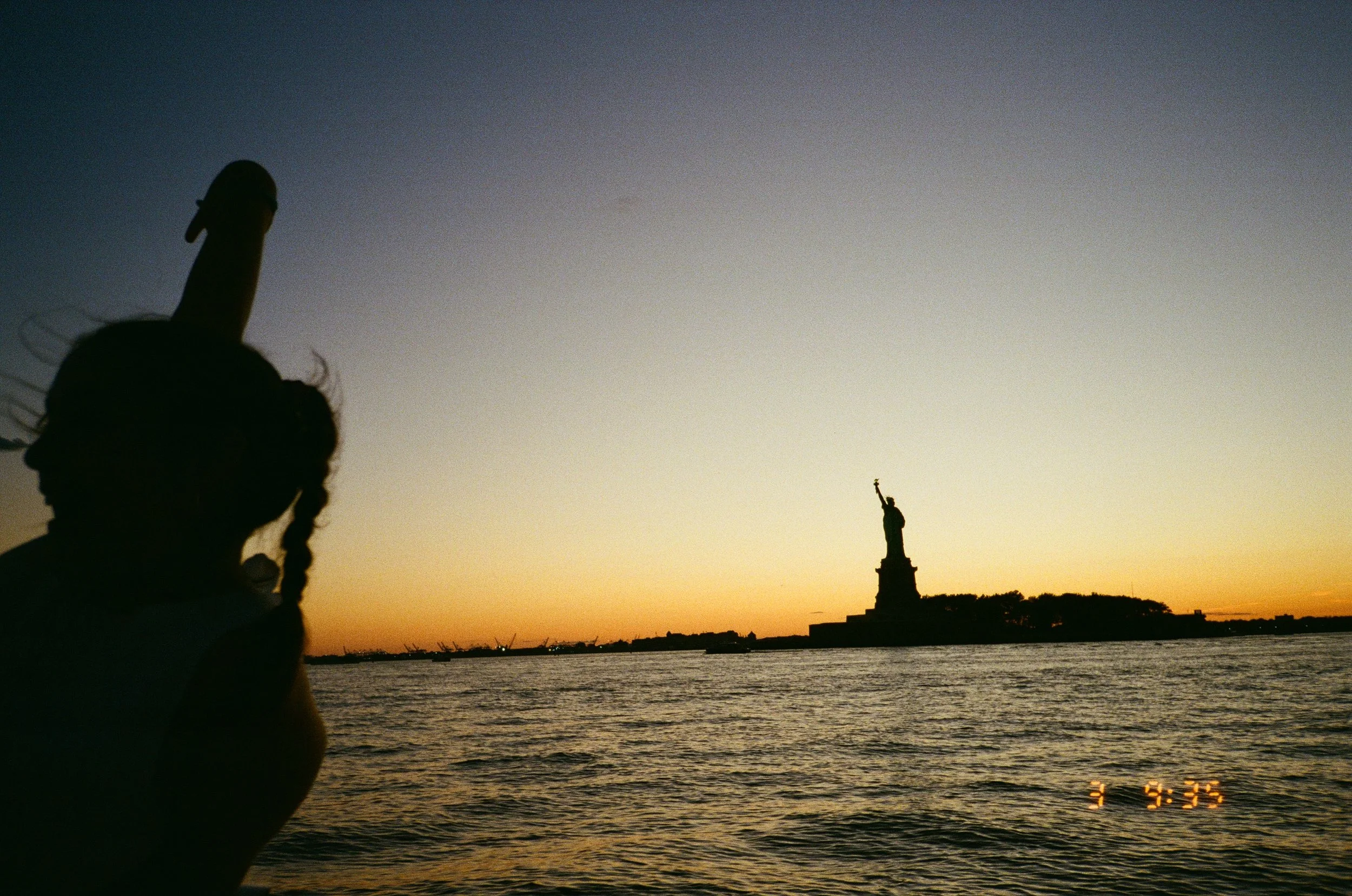 Silhouette of a girl with braided hair and a fedora hat, gesturing with her middle finger, with the Statue of Liberty and the New York City skyline in the background at sunset.
