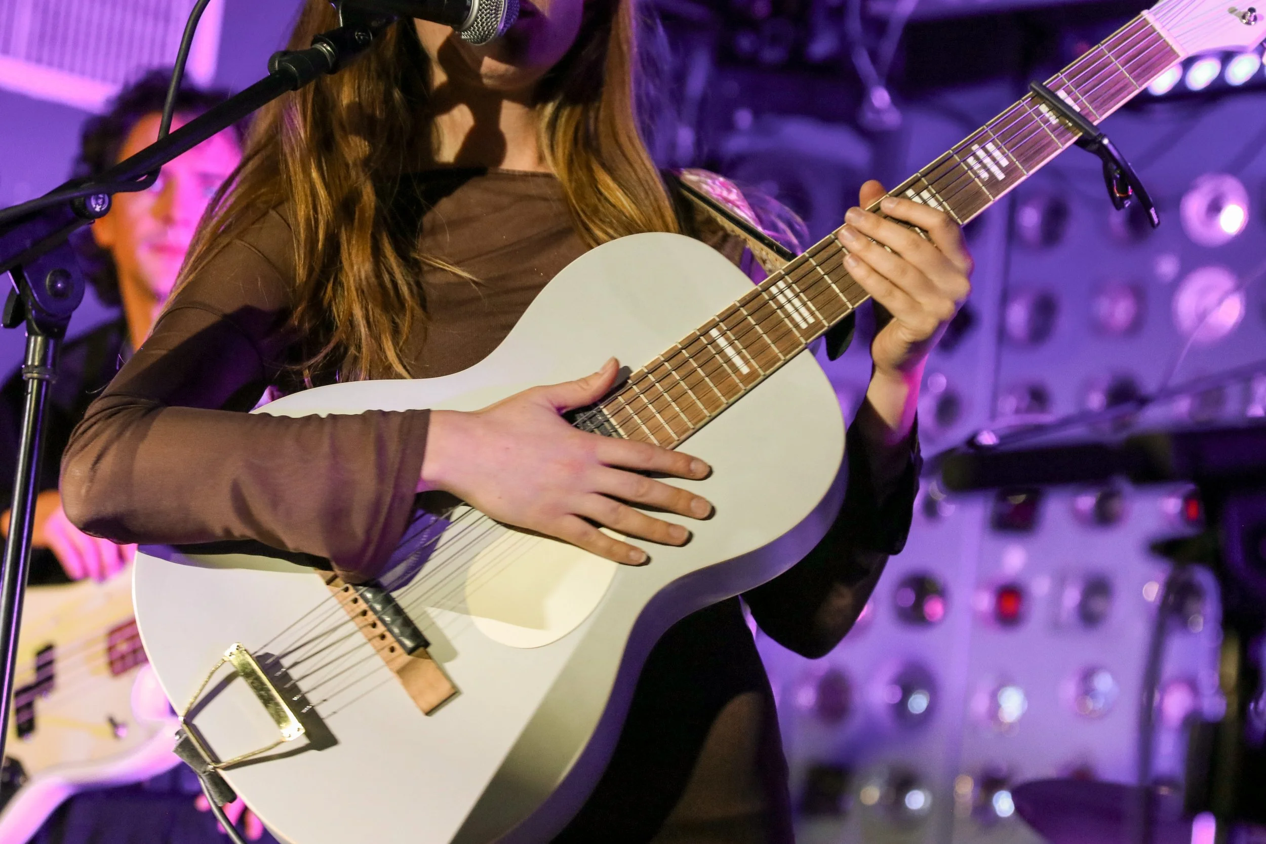 A woman playing an acoustic guitar while singing into a microphone during a live performance, with purple stage lighting in the background.