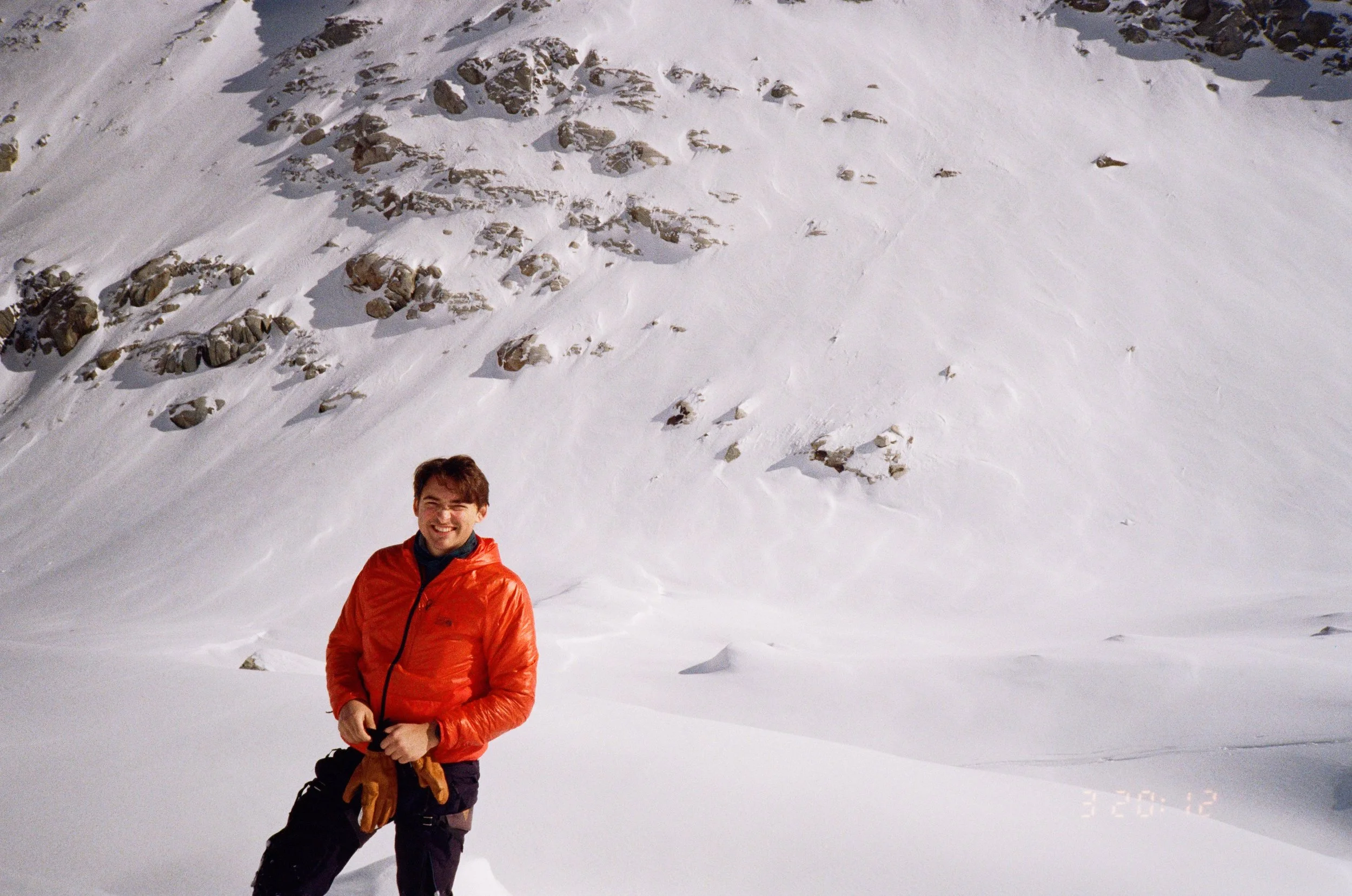 A man smiling, wearing an orange jacket and black pants, standing in snow-covered mountains with rocky slopes in the background.