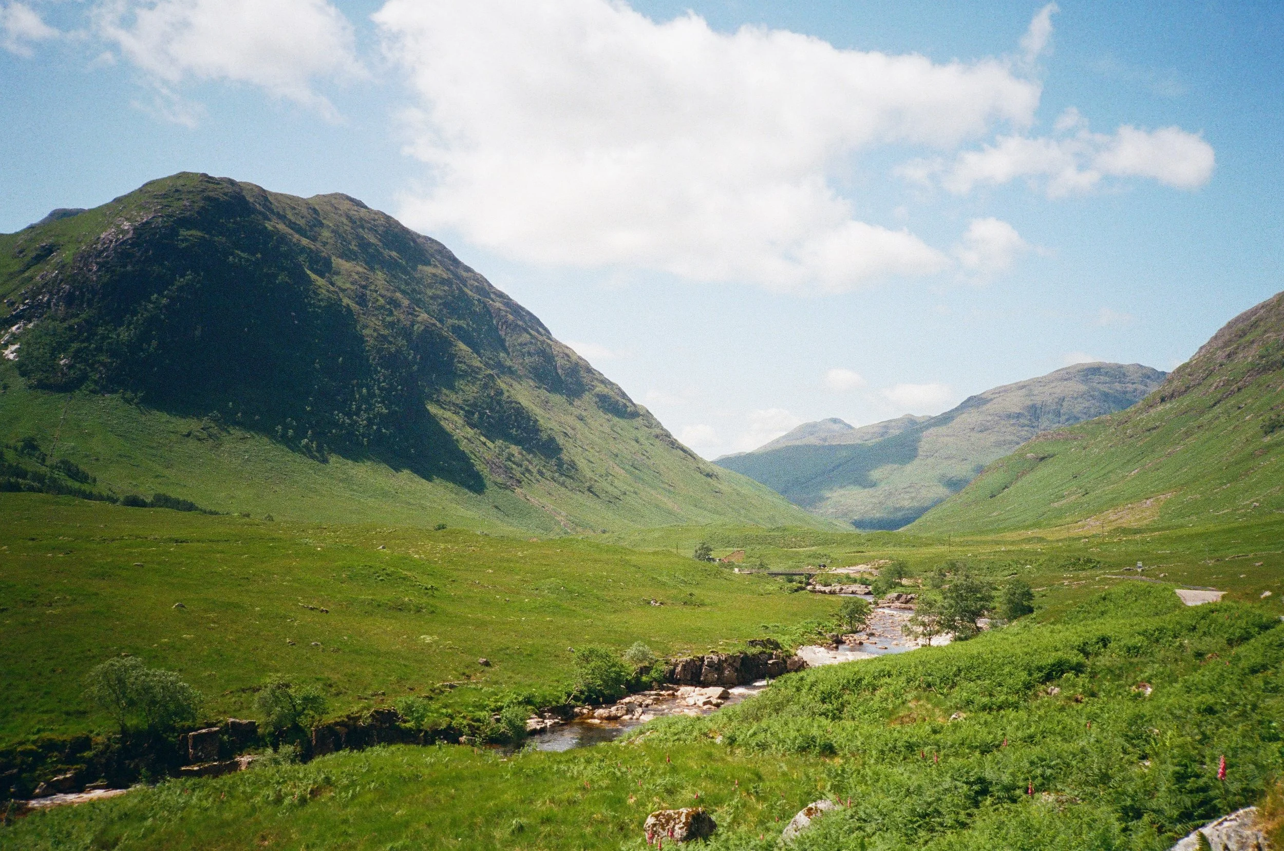 A scenic landscape of rolling green hills and mountains under a partly cloudy sky with a small stream flowing through the valley.