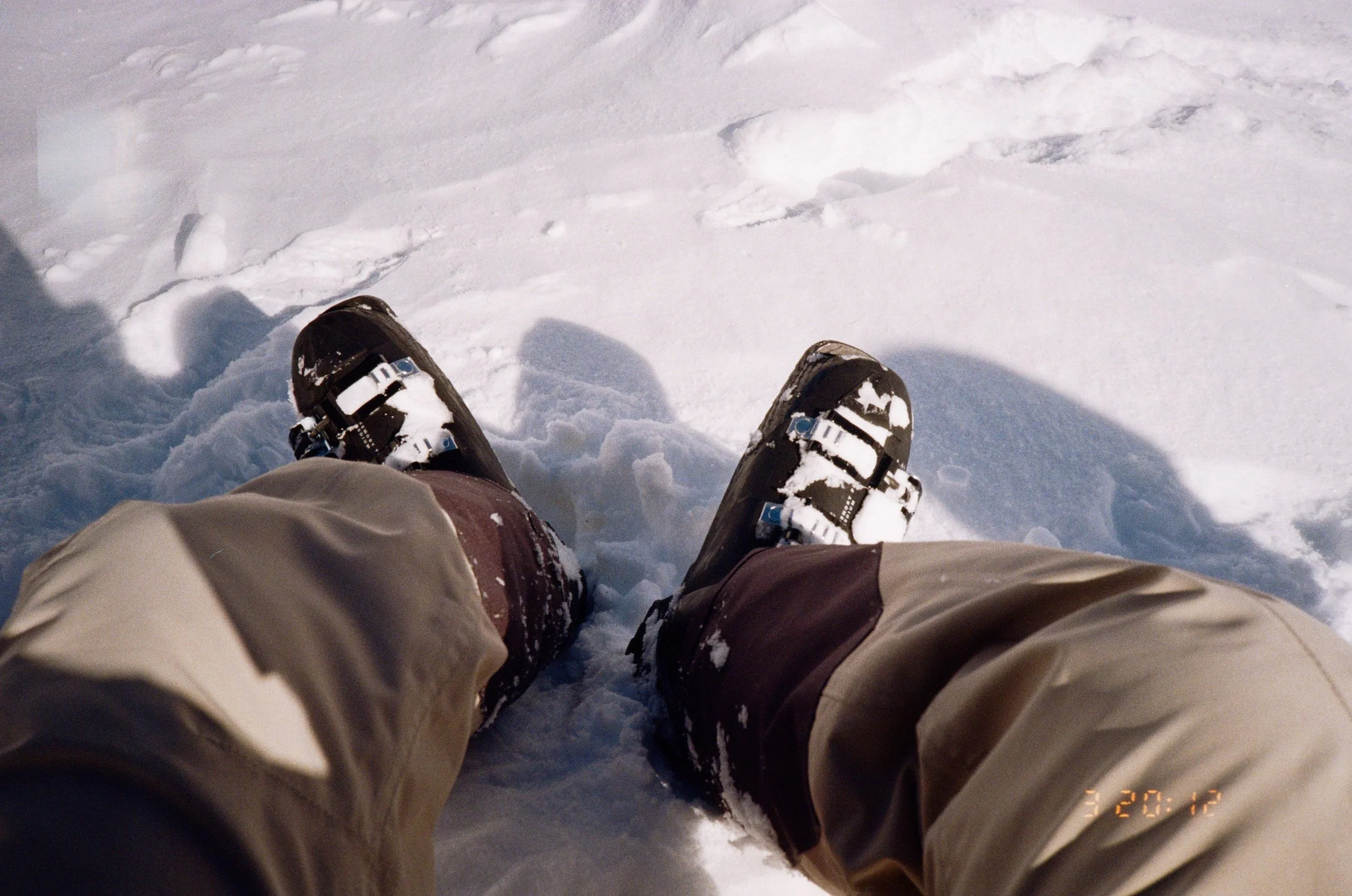 View of someone's legs wearing snow boots and beige pants, sitting in snow-covered ground, with snow and footprints visible.