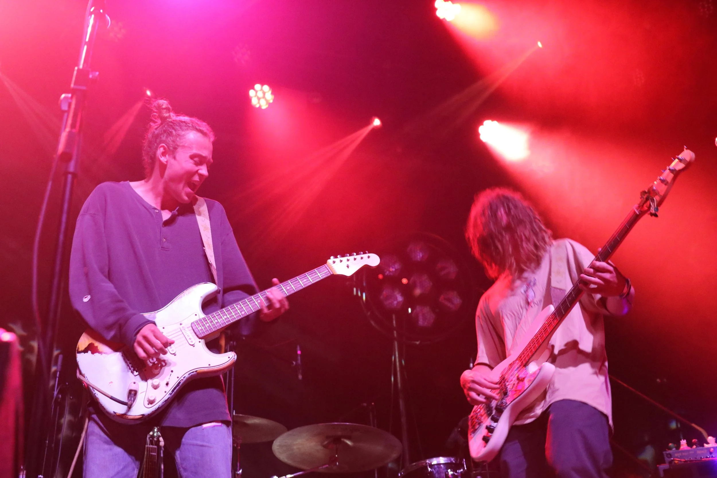 Two musicians perform on stage with guitars under red lighting; one person has curly hair and is playing an electric guitar, the other with long hair playing an electric bass.