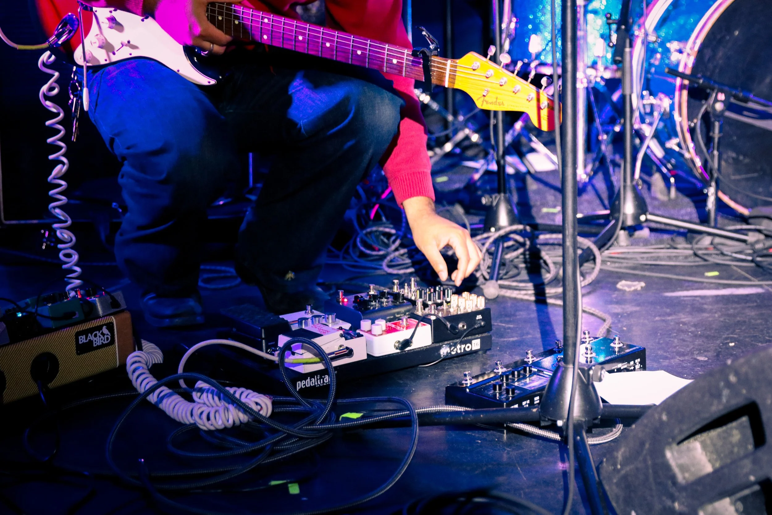 A musician on stage adjusting electronic music equipment with a guitar resting on their lap. Numerous cables and pedals are visible on the floor, and drums are in the background.