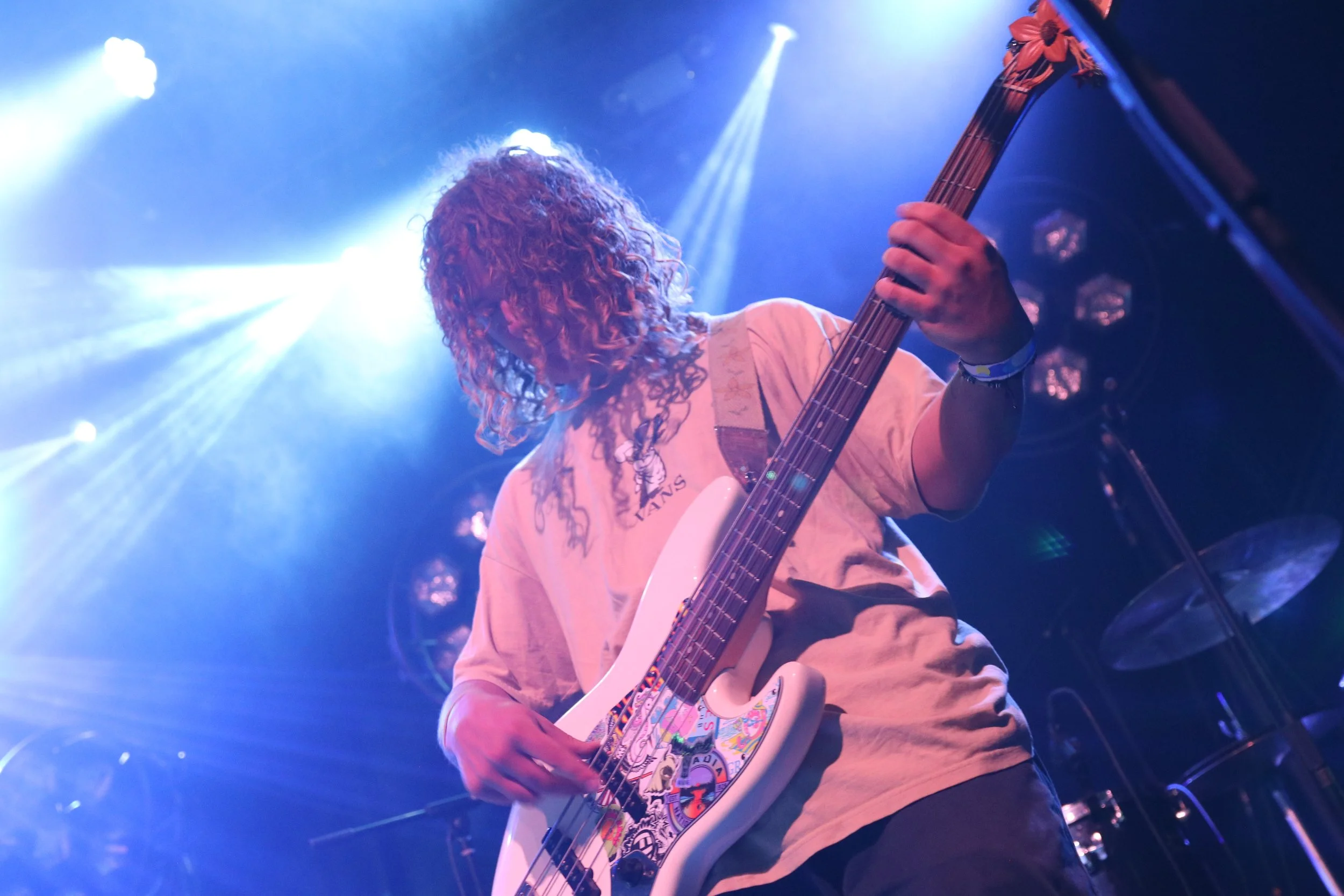 A person playing a white bass guitar with colorful stickers, performing on stage with bright stage lights shining behind them.