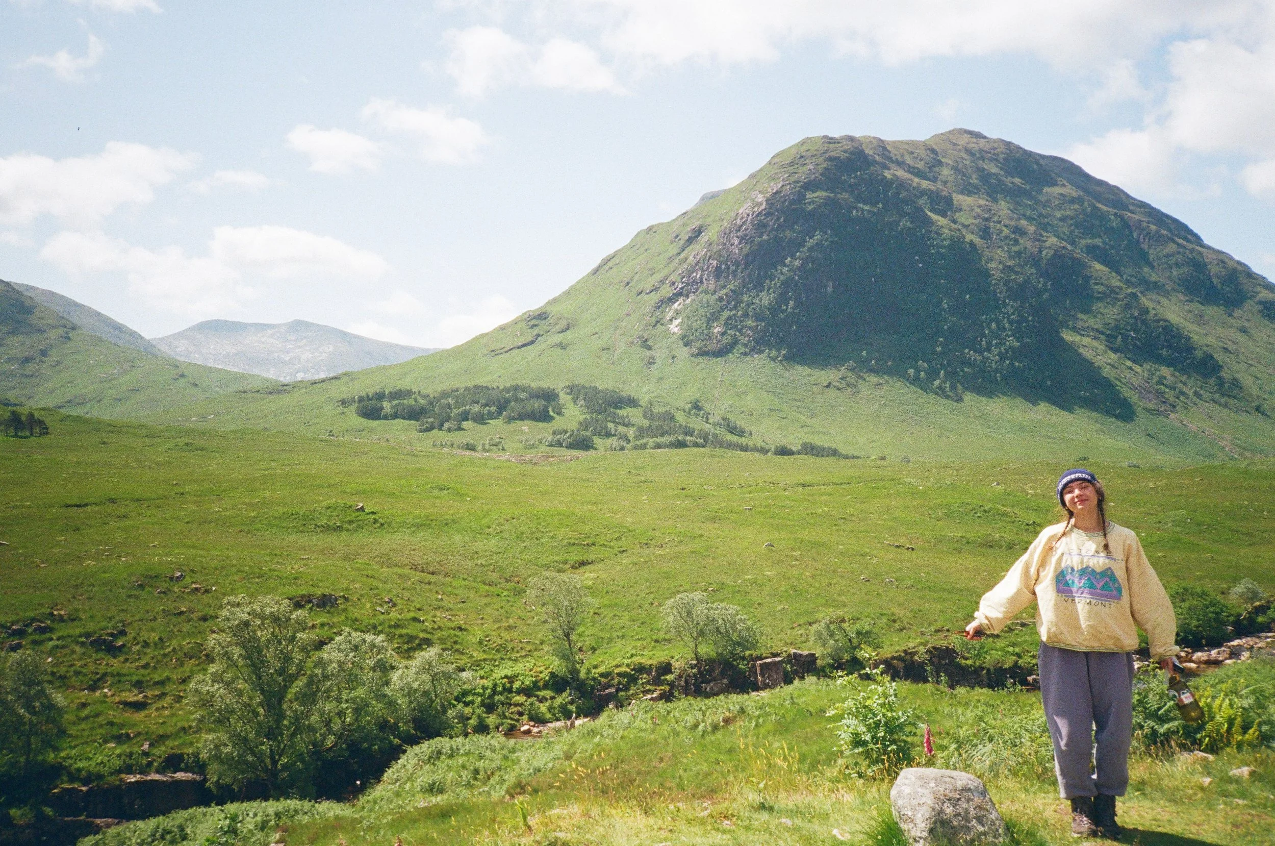 A woman standing in a lush green valley with mountains in the background under a partly cloudy sky, holding a drink in one hand.