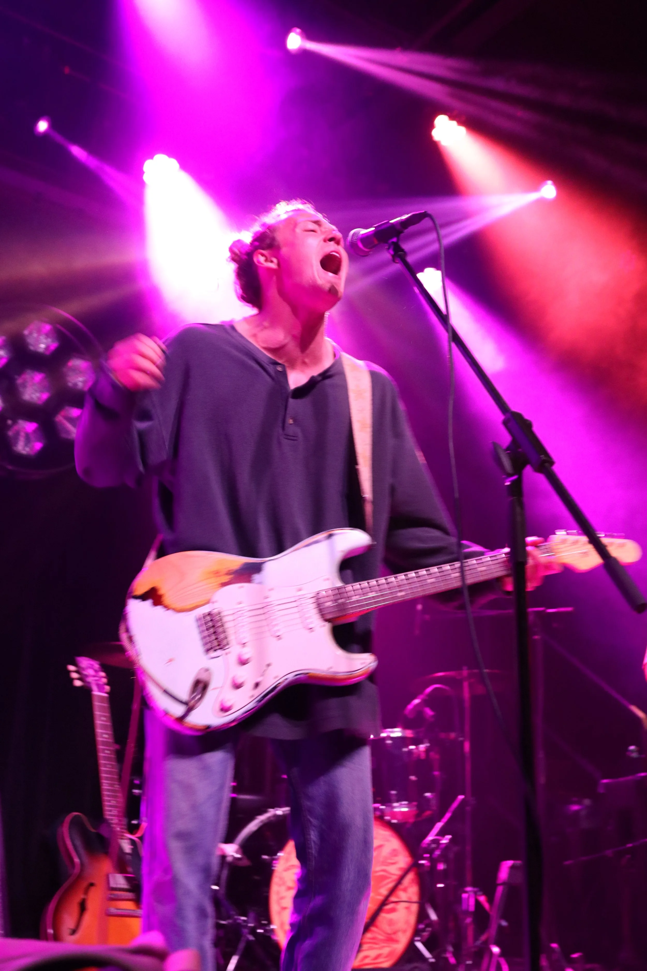 Musician singing passionately into a microphone while playing an electric guitar on stage with purple and pink stage lights.